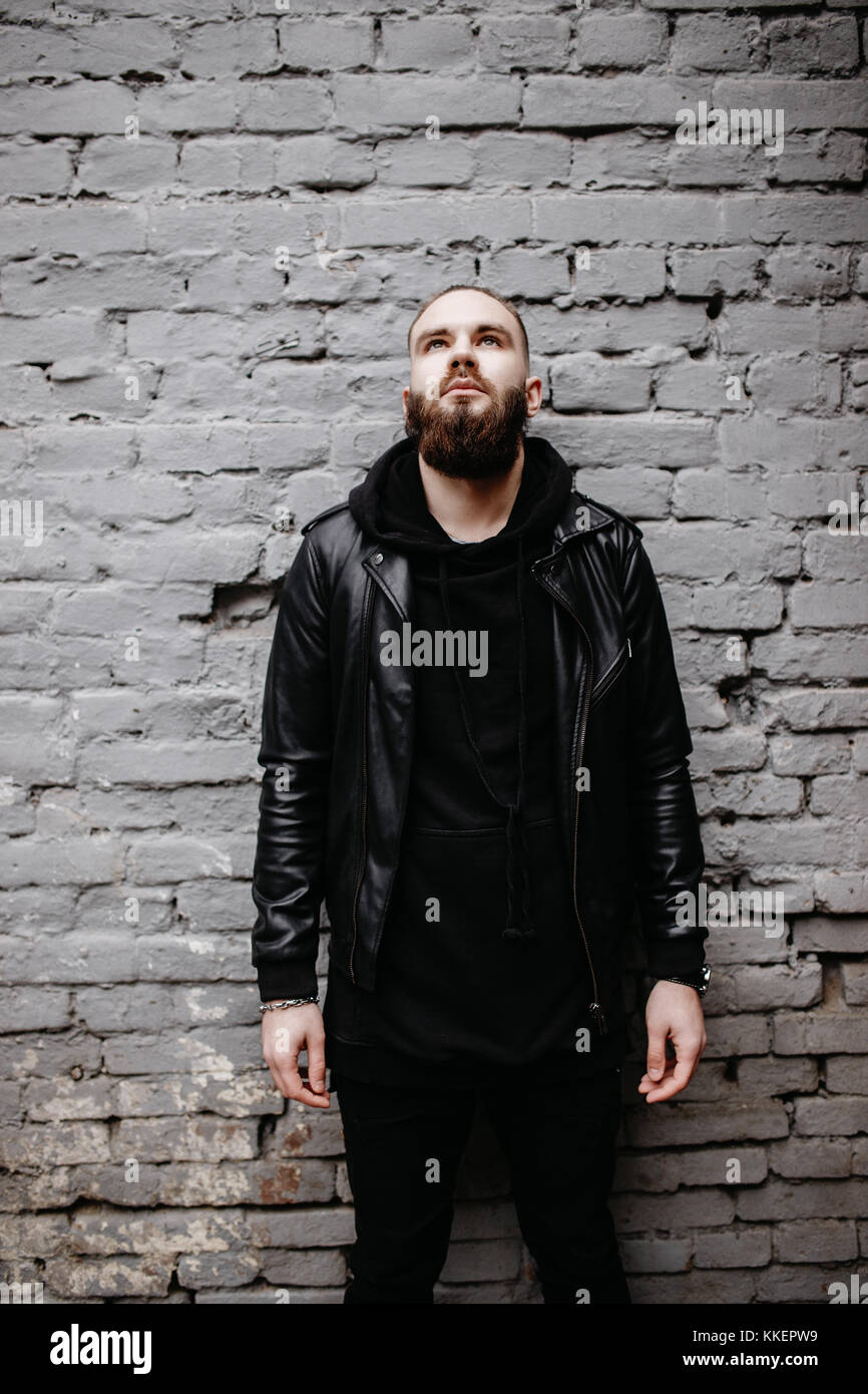Jeune homme barbu moderne en noir vêtements de style posant contre un mur de briques. Banque D'Images