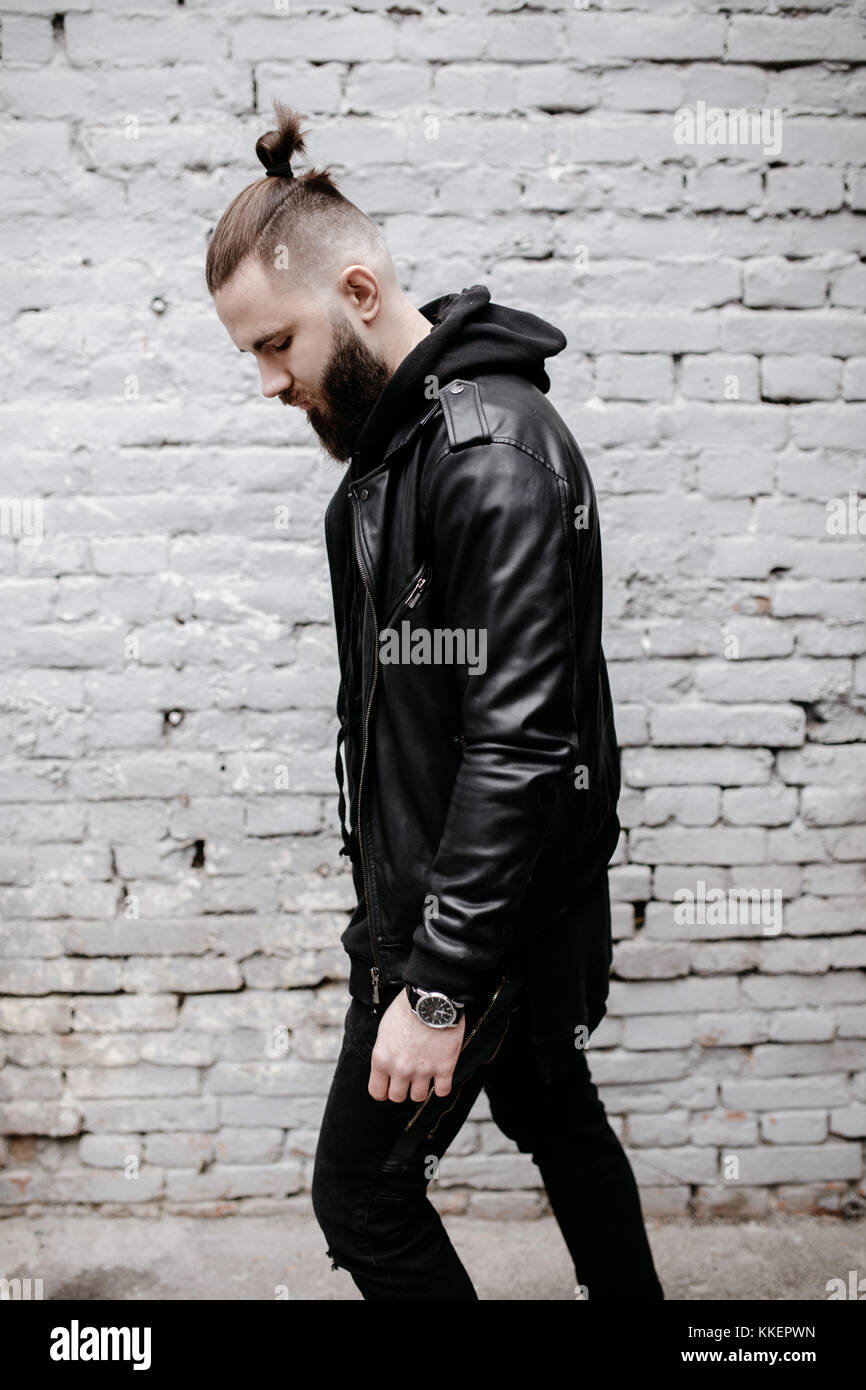 Jeune homme barbu moderne en noir vêtements de style posant contre un mur de briques. Banque D'Images