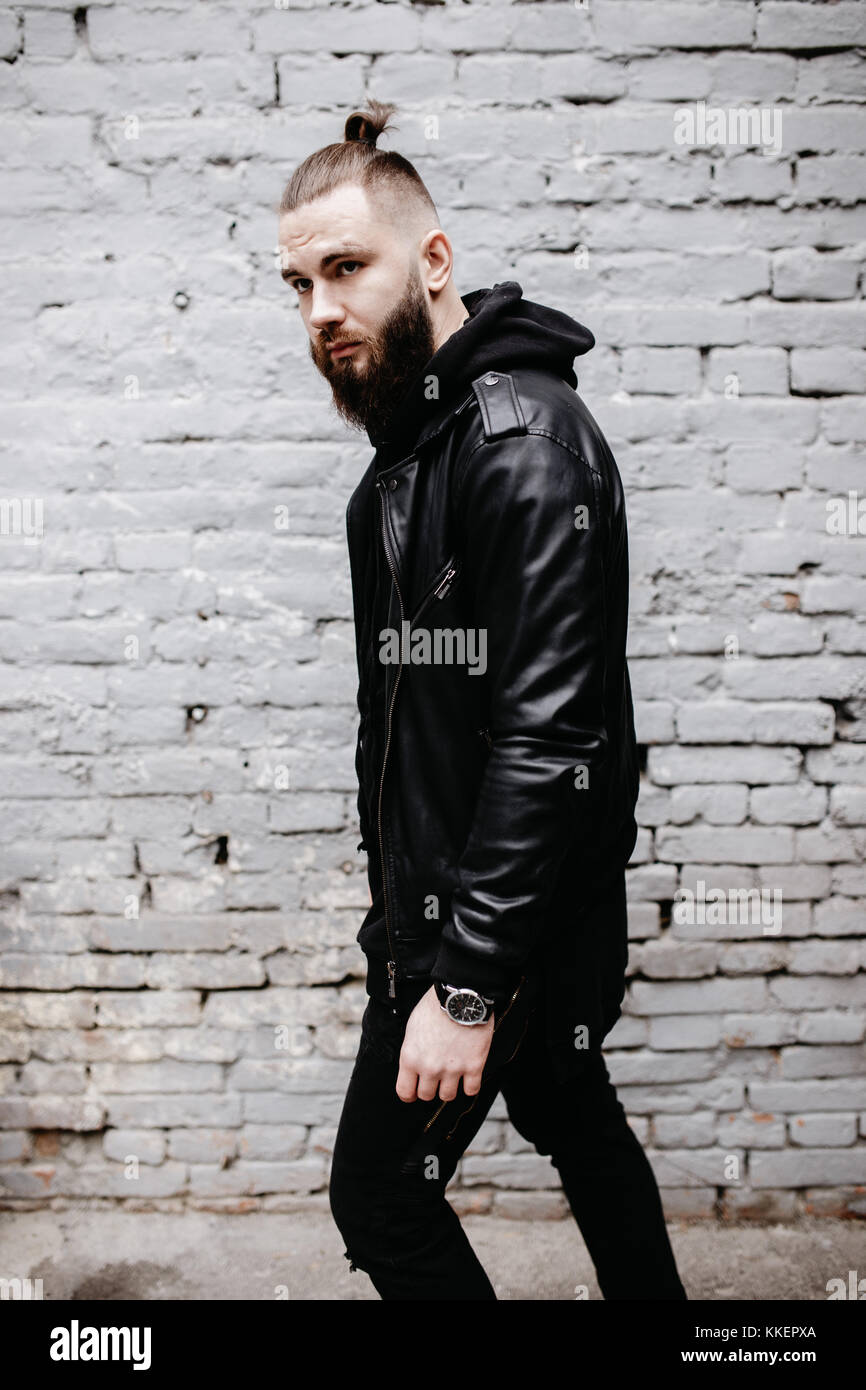 Jeune homme barbu moderne en noir vêtements de style posant contre un mur de briques. Banque D'Images