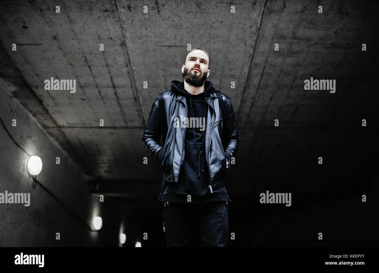 Jeune homme barbu moderne en noir vêtements de style debout dans autour d'origine urbaine. Banque D'Images Jeune homme barbu moderne en noir vêtements de style debout dans autour d'origine urbaine. Banque D'Images