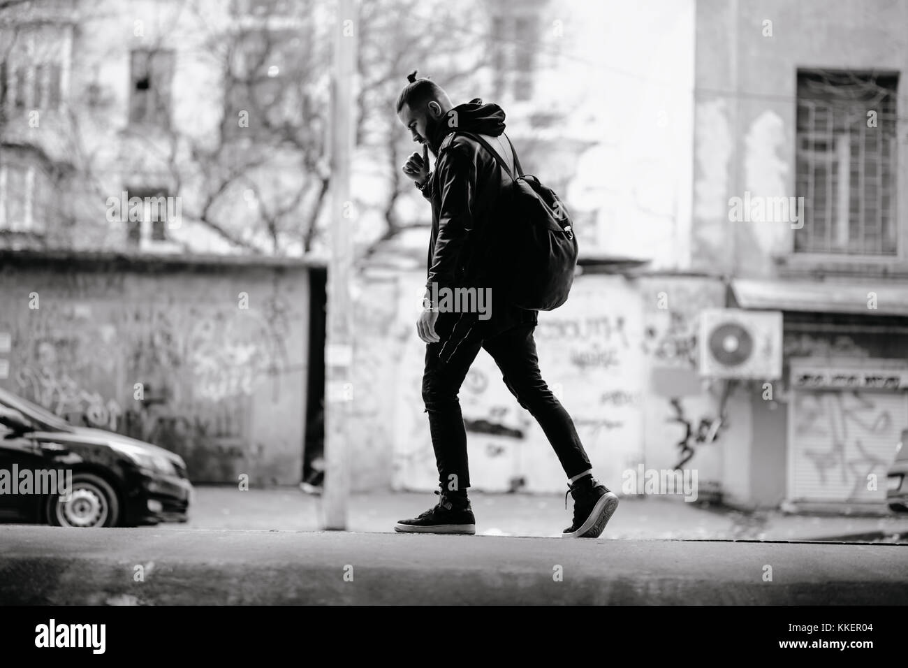 Jeune homme barbu moderne en noir vêtements de style debout dans autour d'origine urbaine. Banque D'Images Jeune homme barbu moderne en noir vêtements de style debout dans autour d'origine urbaine. Banque D'Images