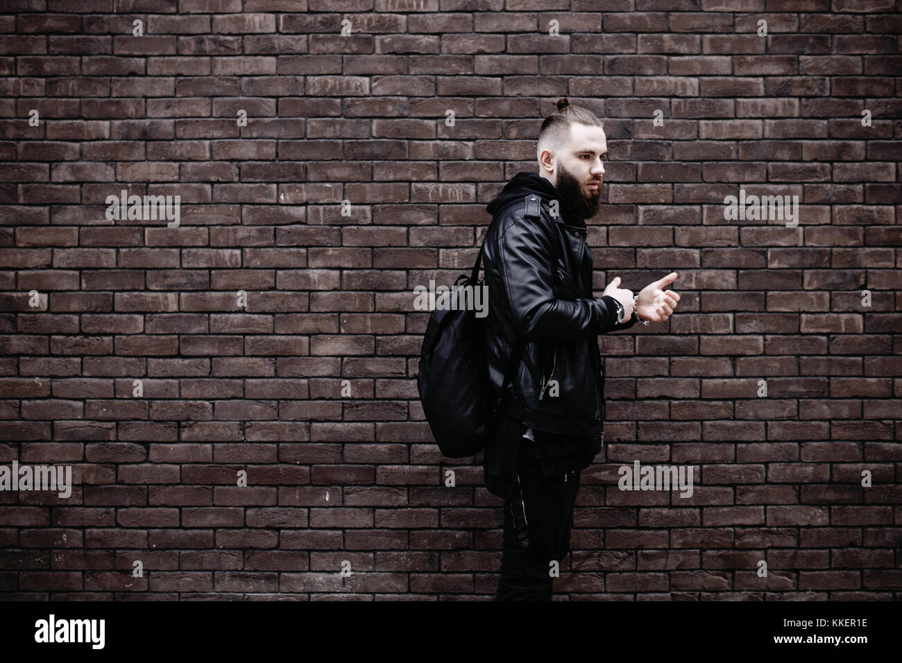 Jeune homme barbu moderne en noir vêtements de style posant contre un mur de briques. Banque D'Images