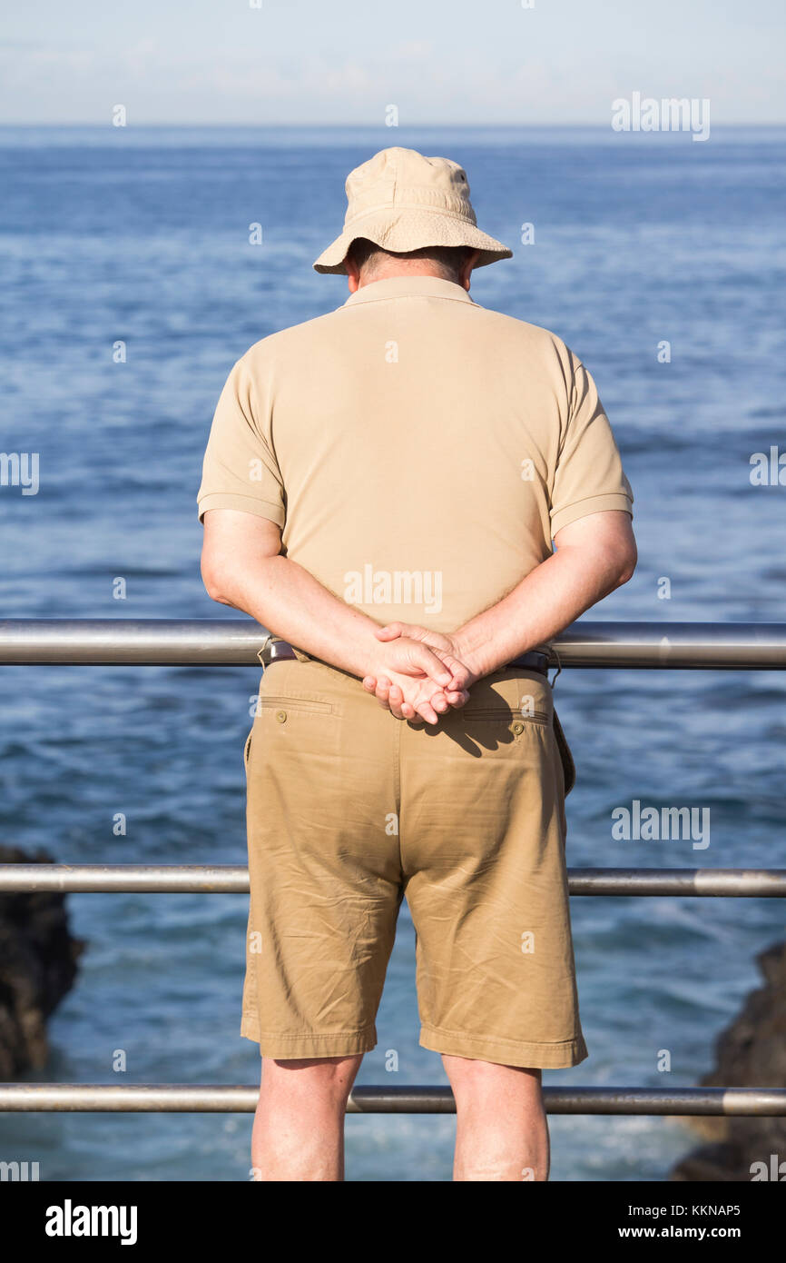 Homme vêtu d'un chapeau, d'une chemise et d'un short beiges qui donne sur la mer. Banque D'Images