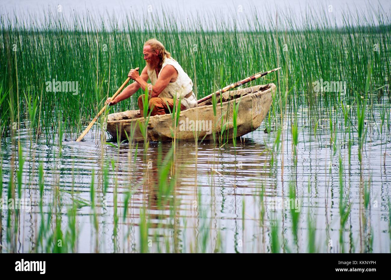 Re-enactment néolithique. L'homme de l'âge de pierre avec foëne paddling peau d'animal sur bateau coracle Reed Lake Shore. Kilmartin, Ecosse Banque D'Images