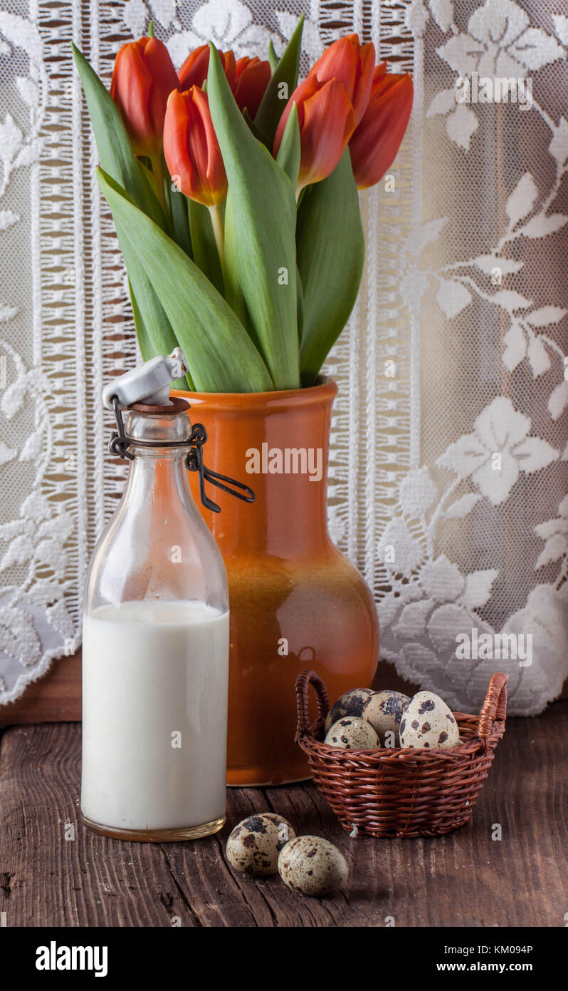 Oeuf de caille en brun panier avec une bouteille de lait et pot en céramique de tulipes rouges sur la vieille table en bois Banque D'Images