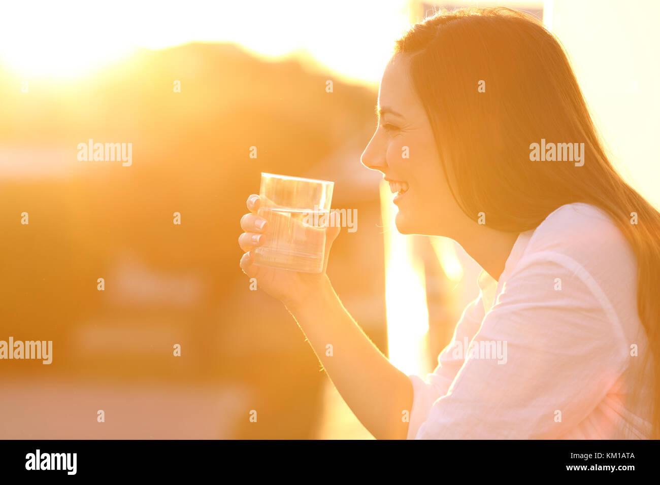 Side view portrait of a happy dame tenant un verre d'eau à l'extérieur dans un balcon au coucher du soleil Banque D'Images