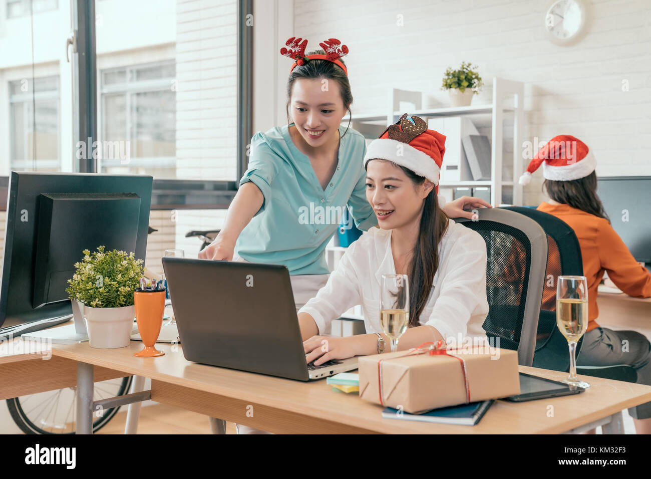 Deux femmes d'affaires heureux de vérifier en ligne vente de Noël tandis que leur collègue travaillant dans l'arrière-plan. Banque D'Images Deux femmes d'affaires heureux de vérifier en ligne vente de Noël tandis que leur collègue travaillant dans l'arrière-plan. Banque D'Images
