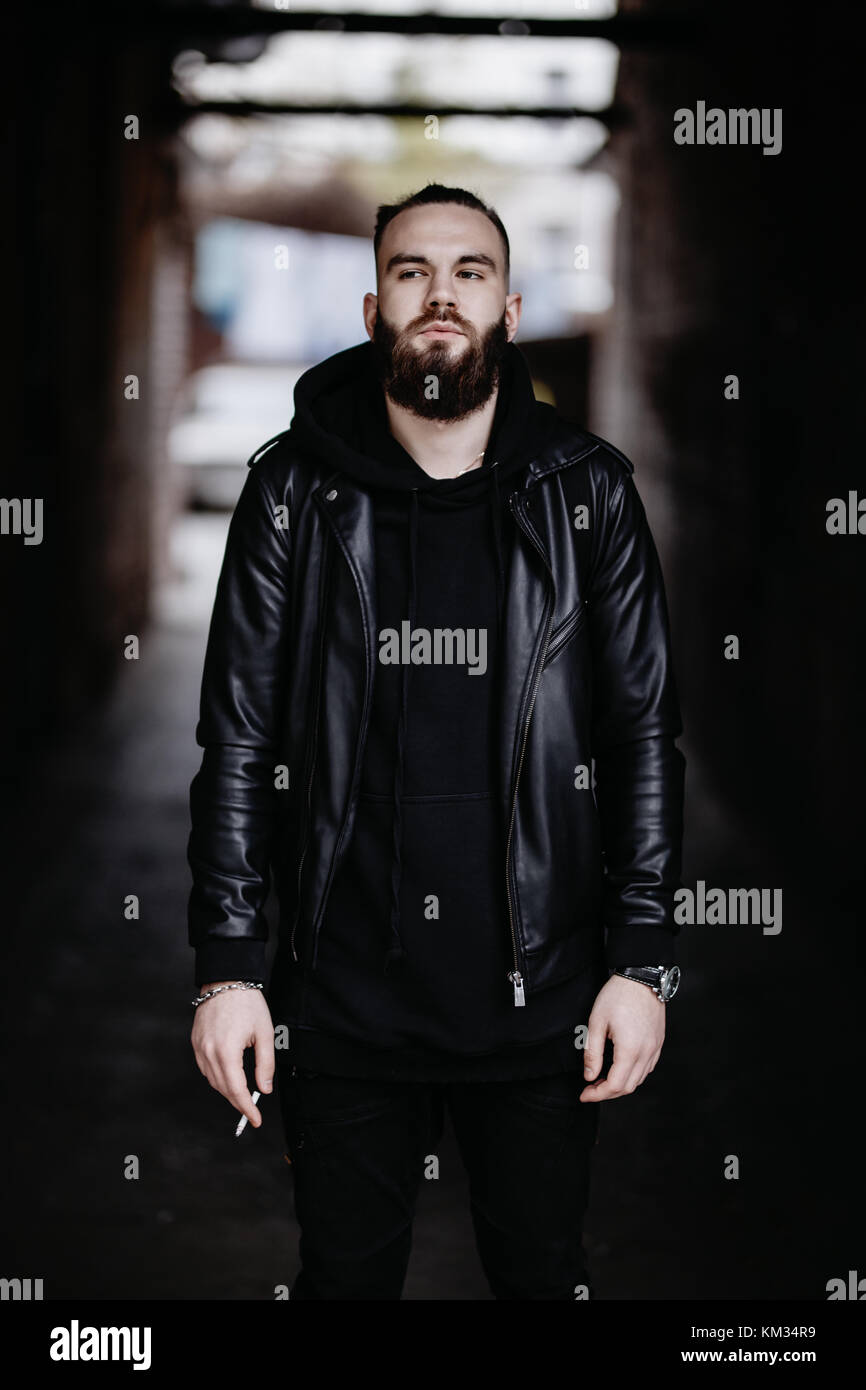 Jeune homme barbu moderne en noir vêtements de style l'article en contexte urbain. Banque D'Images Jeune homme barbu moderne en noir vêtements de style l'article en contexte urbain. Banque D'Images