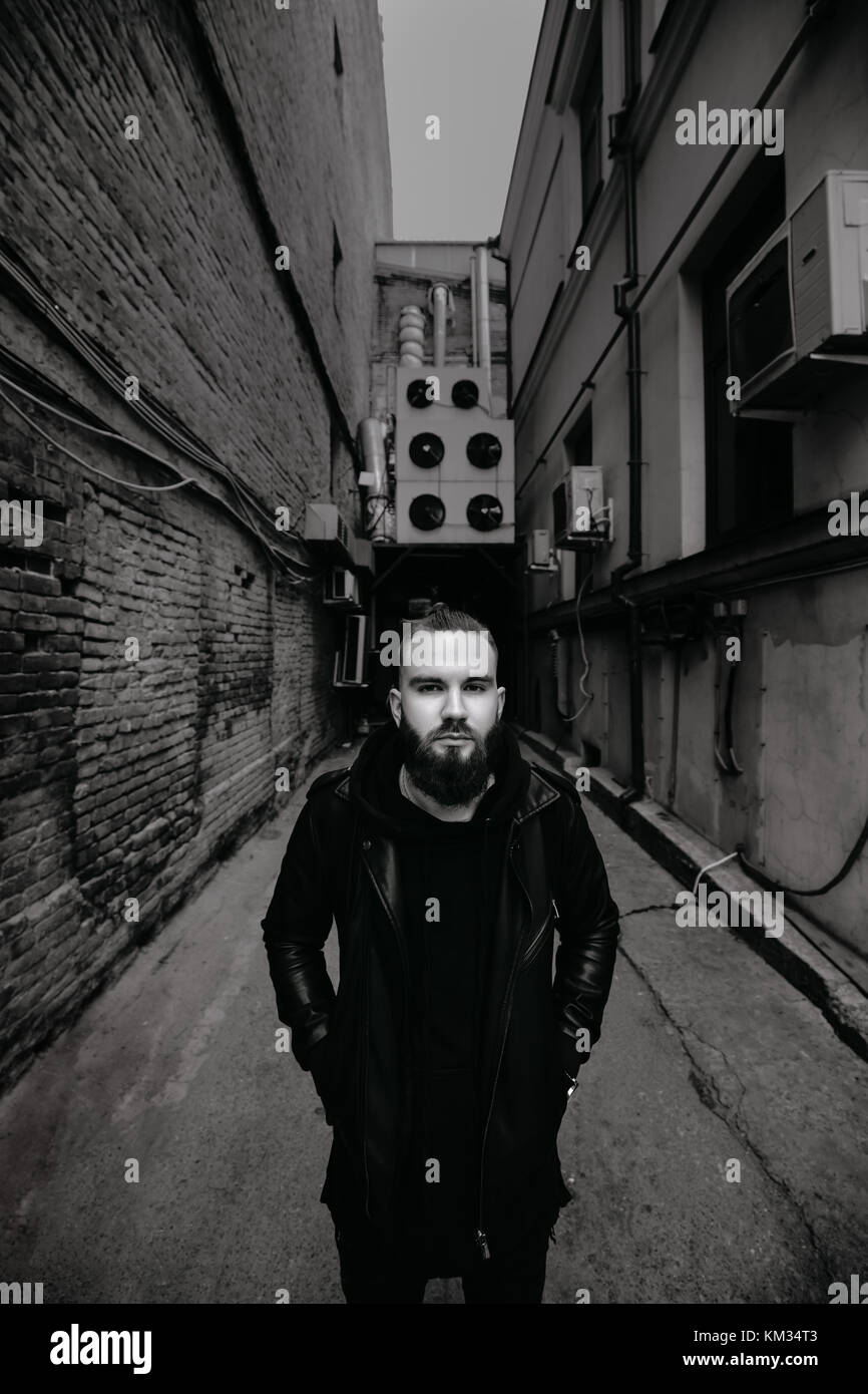 Jeune homme barbu moderne en noir vêtements de style l'article en contexte urbain. Banque D'Images Jeune homme barbu moderne en noir vêtements de style l'article en contexte urbain. Banque D'Images