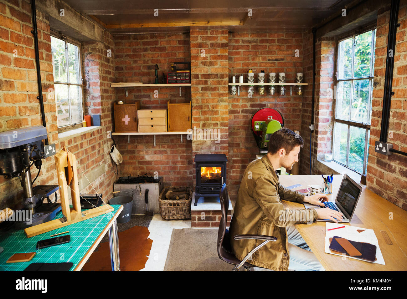 Un designer assis dans son atelier, le travail du cuir à un bureau à l'aide d'un ordinateur portable. Poêle à bois avec un feu allumé. Banque D'Images