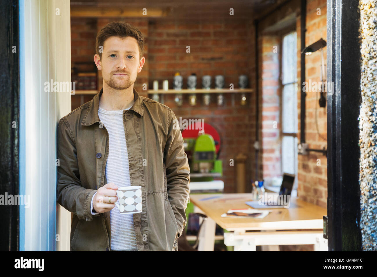 Un jeune homme debout à un atelier porte, tenant une tasse de café. Banque D'Images