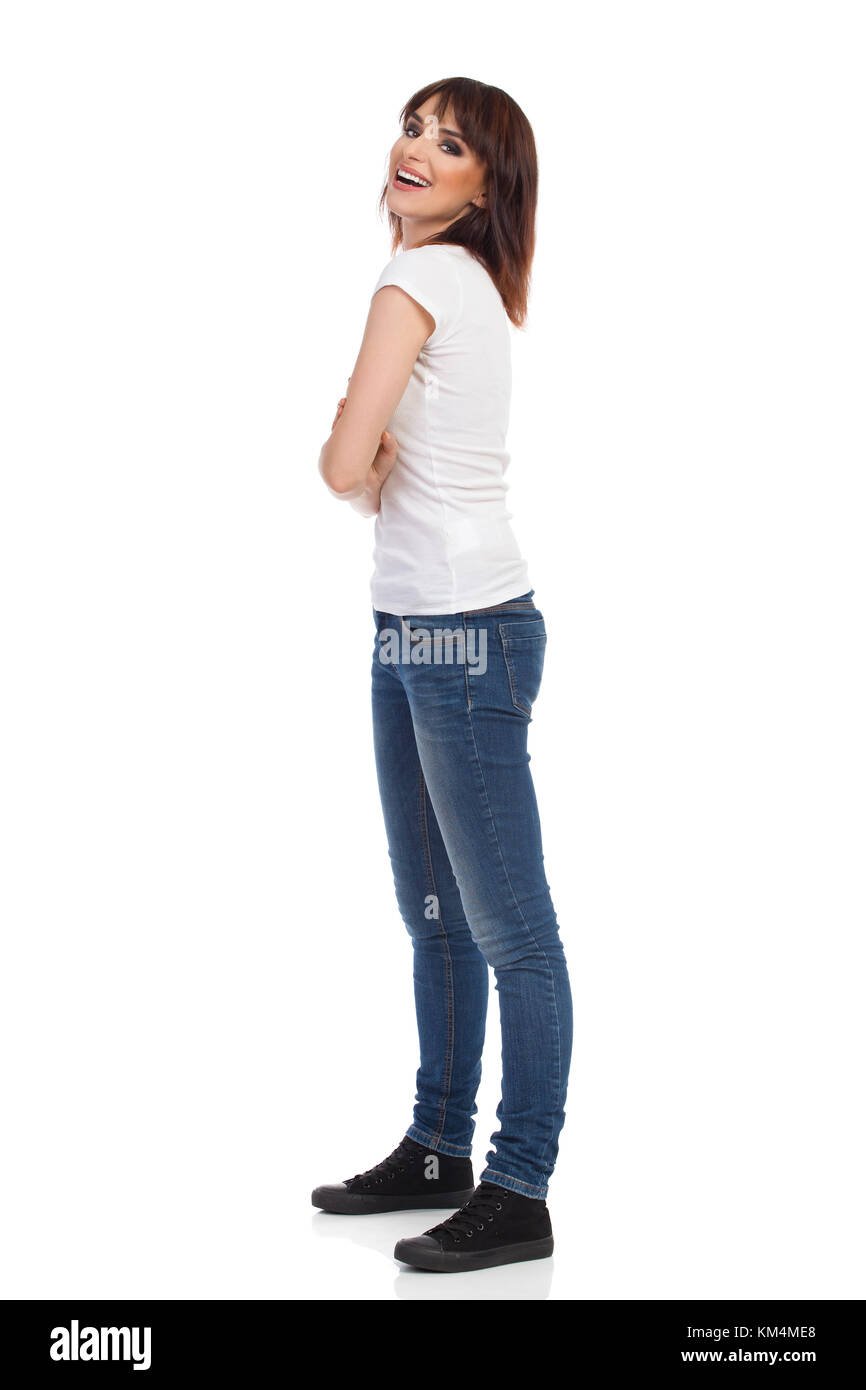 Jeune femme en jeans et t-shirt blanc est debout avec rms croisés, looking at camera and smiling. full length portrait isolé sur blanc. Banque D'Images