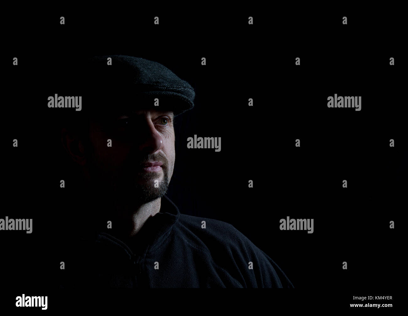 Portrait photo bas-clé d'homme barbu en casquette sombre, face à demi-lumière, pose pensive. Eclairage faible, fond noir : effet chiaroscuro artistique. Banque D'Images
