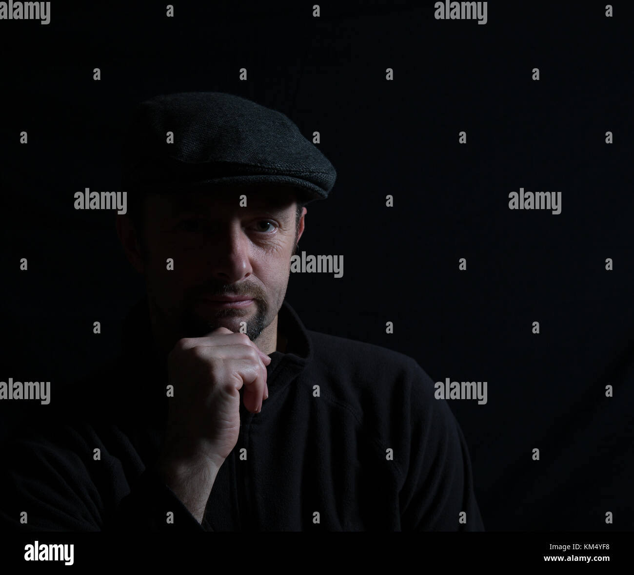 Portrait artistique d'un homme isolé en casquette noire, face à demi-lumière, affamé à l'appareil photo, main sur le menton, sous les projecteurs. Un éclairage subtil crée une image ombragée. Banque D'Images