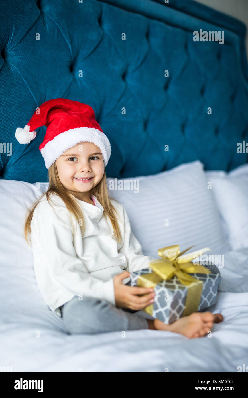 Heureux sort little girl in santa hat holding gift box, looking at camera while sitting on bed Banque D'Images