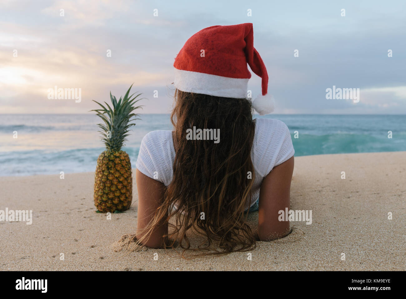 Femme portant un chapeau de Noël allongé sur la plage à côté d'un ananas, Haleiwa, Hawaii, États-Unis Banque D'Images