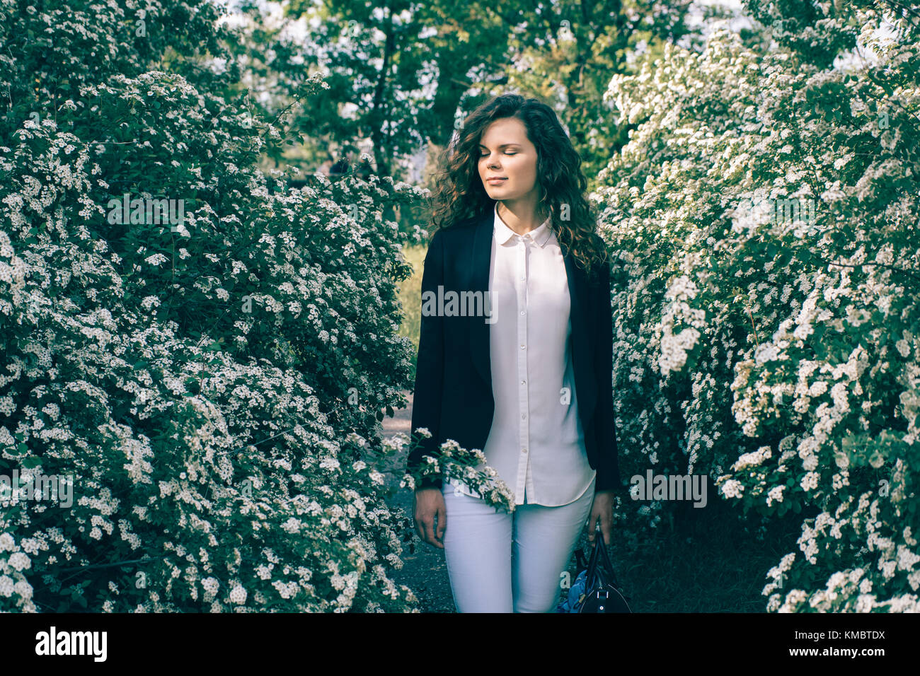 Belle jeune femme en une veste et un jean se trouve dans un jardin parmi les fleurs blanches Banque D'Images