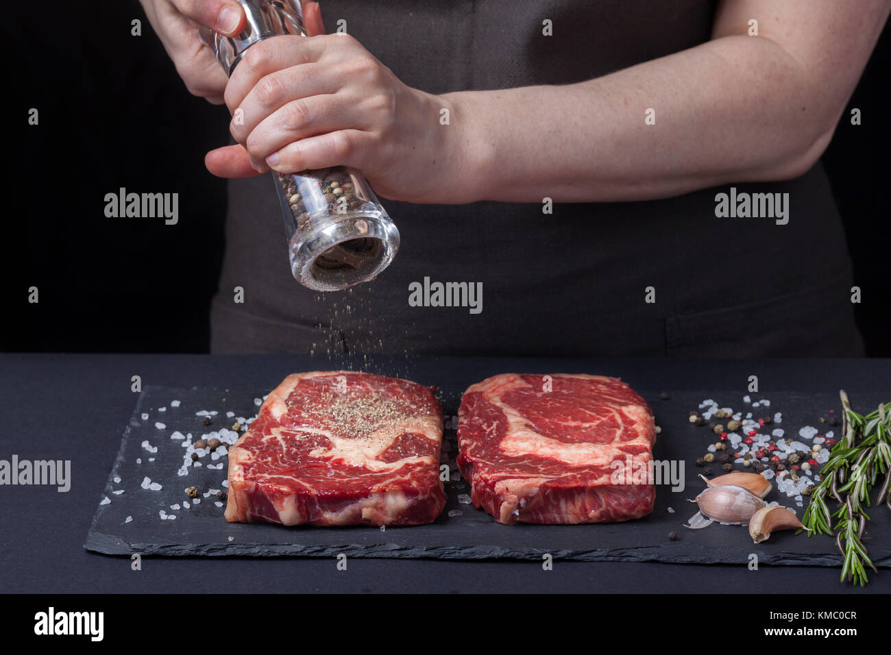 Une femme cuit avec des poivrons deux steaks de ribeye frais crus de bœuf marbré sur un fond sombre.À proximité se trouve un mélange de poivrons, de sel de mer, d'ail et de roo Banque D'Images Une femme cuit avec des poivrons deux steaks de ribeye frais crus de bœuf marbré sur un fond sombre.À proximité se trouve un mélange de poivrons, de sel de mer, d'ail et de roo Banque D'Images