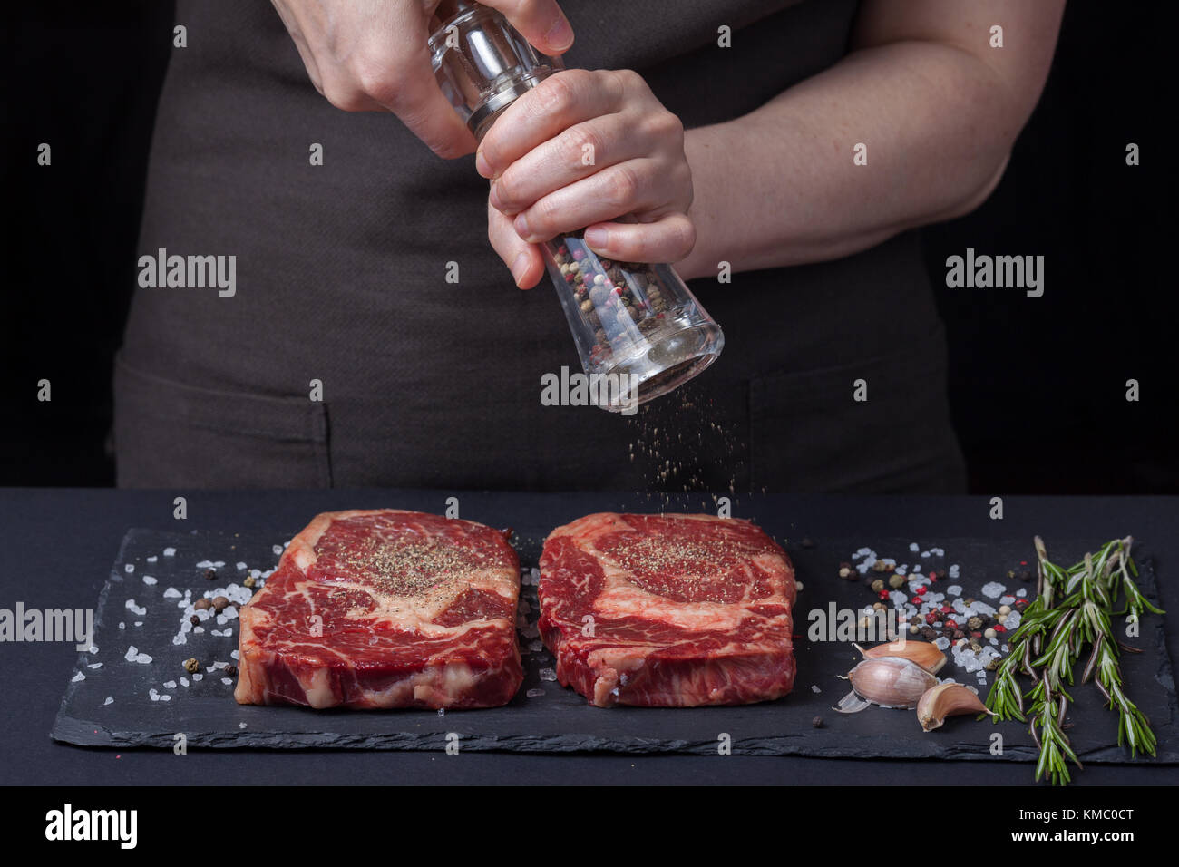 Une femme cuit avec des poivrons deux steaks de ribeye frais crus de bœuf marbré sur un fond sombre.À proximité se trouve un mélange de poivrons, de sel de mer, d'ail et de roo Banque D'Images Une femme cuit avec des poivrons deux steaks de ribeye frais crus de bœuf marbré sur un fond sombre.À proximité se trouve un mélange de poivrons, de sel de mer, d'ail et de roo Banque D'Images