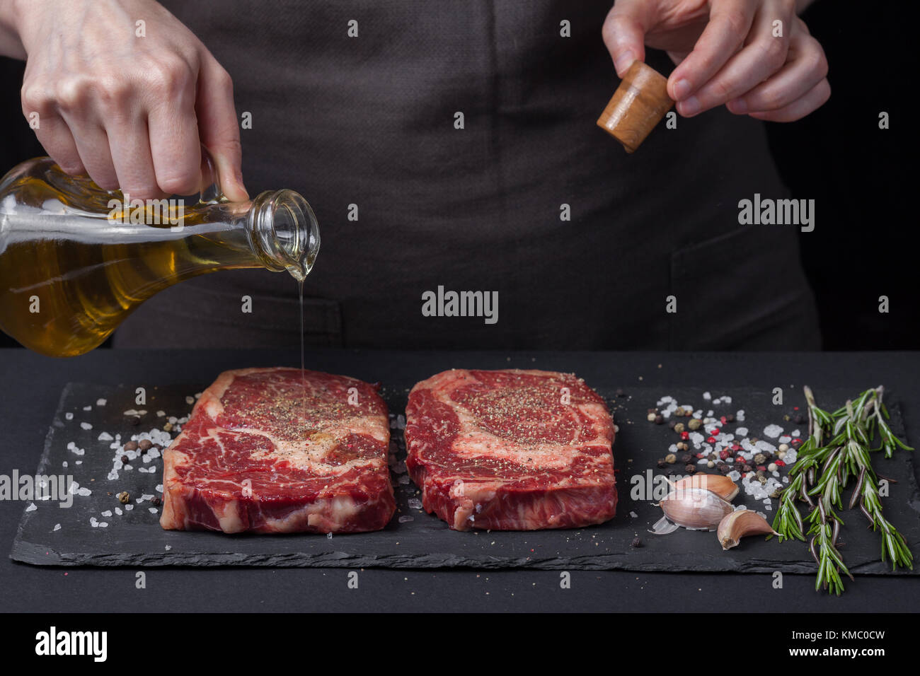 Chef cuisinier qui verse de l'huile d'olive, deux steaks de bœuf cru frais sur fond sombre.À proximité se trouve un mélange de poivrons, de sel de mer, d'ail et de romarin. Banque D'Images Chef cuisinier qui verse de l'huile d'olive, deux steaks de bœuf cru frais sur fond sombre.À proximité se trouve un mélange de poivrons, de sel de mer, d'ail et de romarin. Banque D'Images
