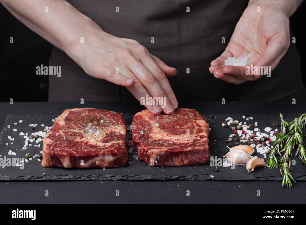 L'assaisonnement féminin du chef frotte deux viandes de bœuf cru sur un fond sombre.À proximité se trouve un mélange de poivrons, de sel de mer, d'ail et de romarin. Banque D'Images L'assaisonnement féminin du chef frotte deux viandes de bœuf cru sur un fond sombre.À proximité se trouve un mélange de poivrons, de sel de mer, d'ail et de romarin. Banque D'Images