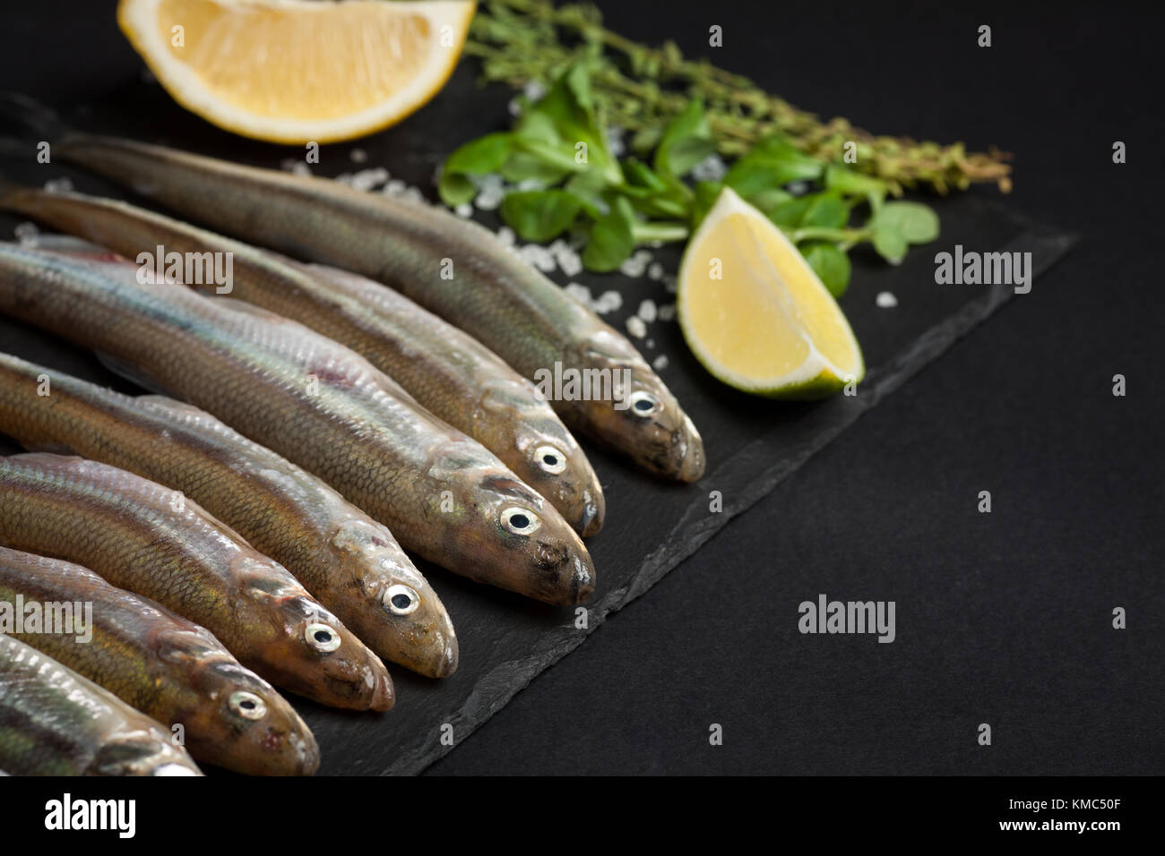 Poisson frais de la mer ou l'éperlan et prête pour la cuisson des sardines au citron, thym, gros sel de mer et sur un fond noir. Le concept de frais, sain de fruits de mer. Vue de dessus Banque D'Images