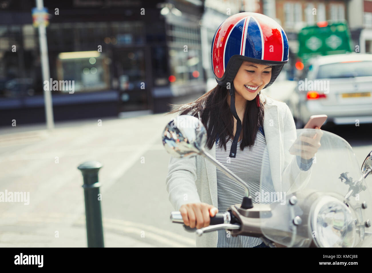 Jeune femme souriante envoyant un SMS avec un téléphone portable sur un scooter à moteur, portant un casque dans la rue urbaine Banque D'Images