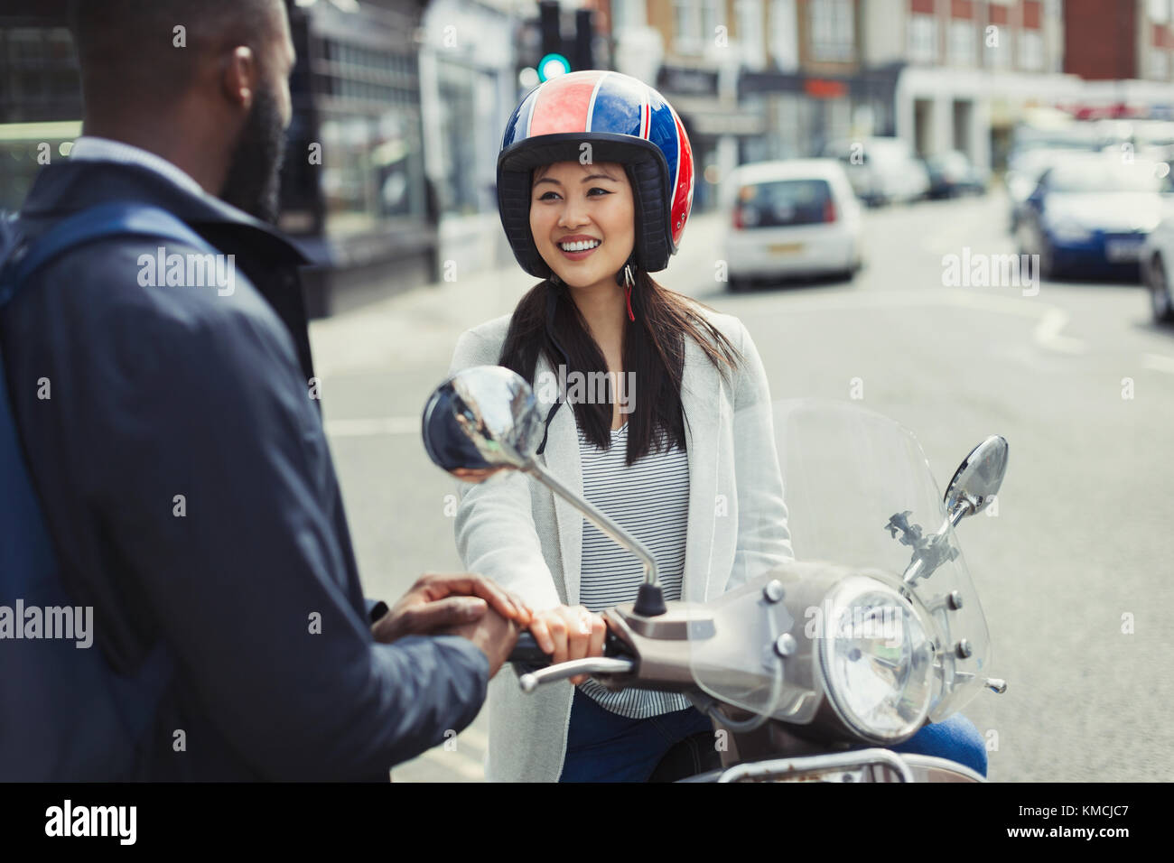 Jeune femme souriante sur scooter de moteur parlant à un ami sur rue urbaine ensoleillée Banque D'Images