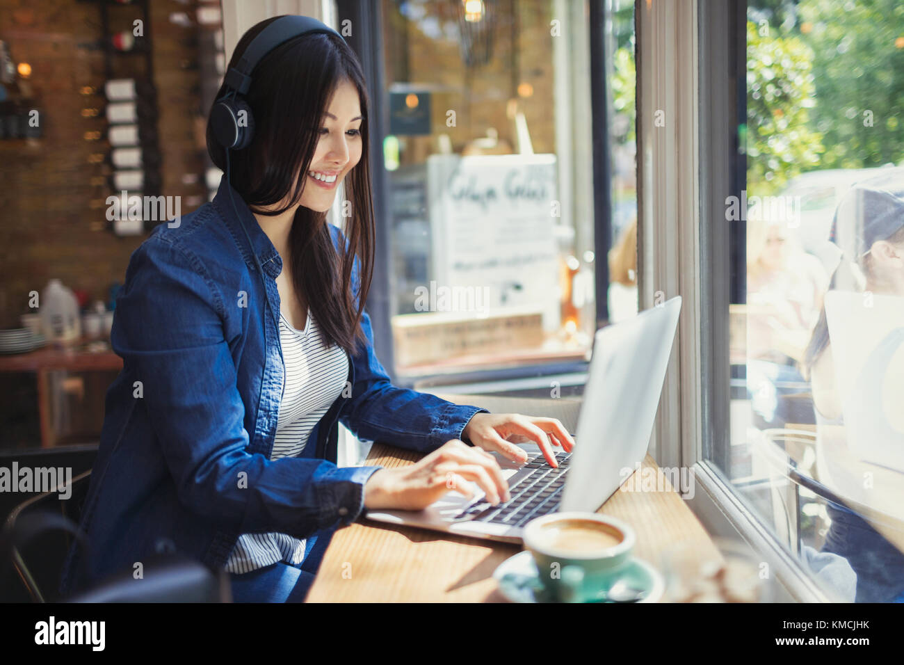 Jeune femme avec casque utilisant un ordinateur portable à la fenêtre ensoleillée du café Banque D'Images