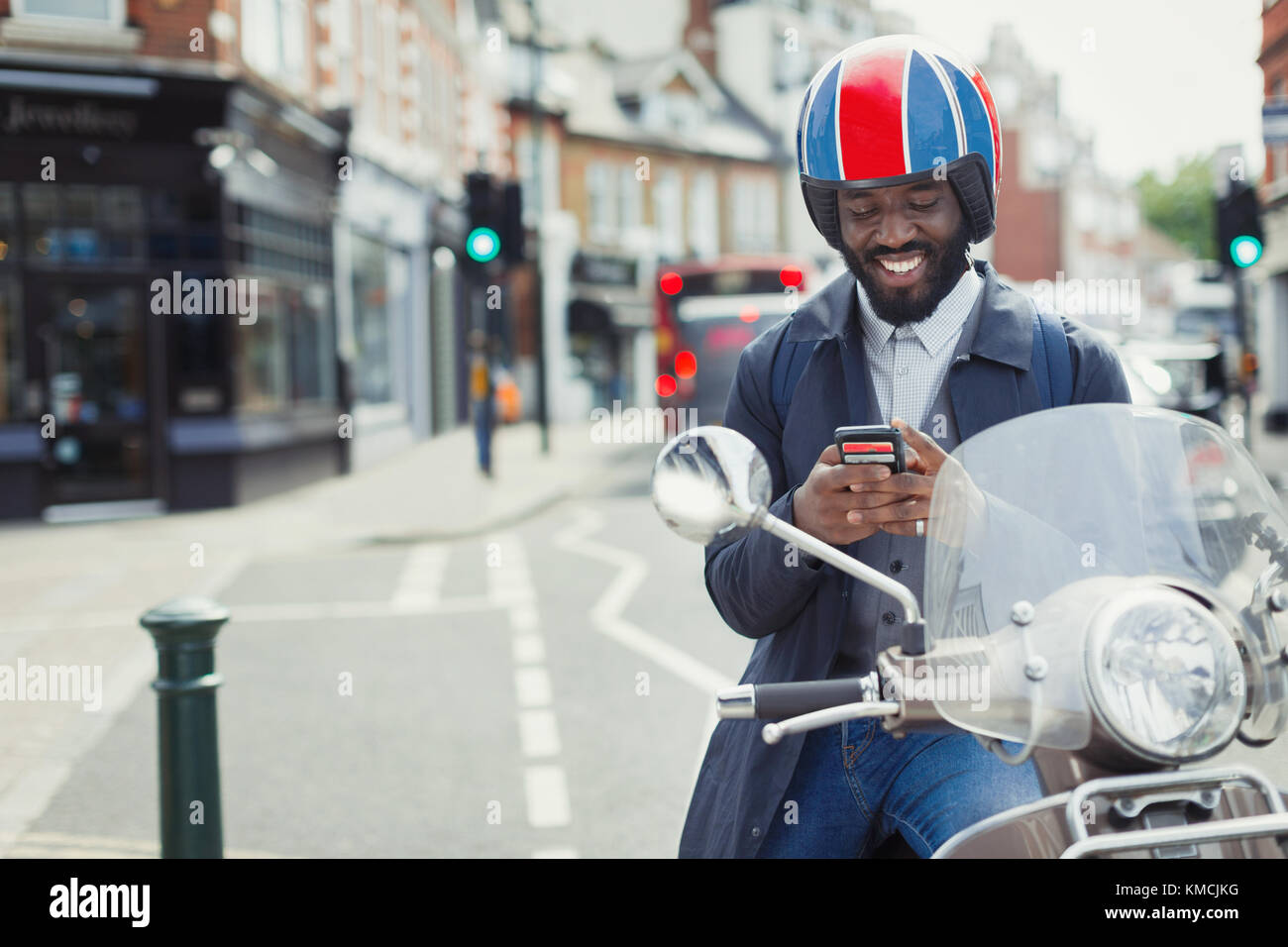 Souriant jeune homme d'affaires dans un casque sur scooter de moteur texting avec téléphone cellulaire dans la rue urbaine Banque D'Images