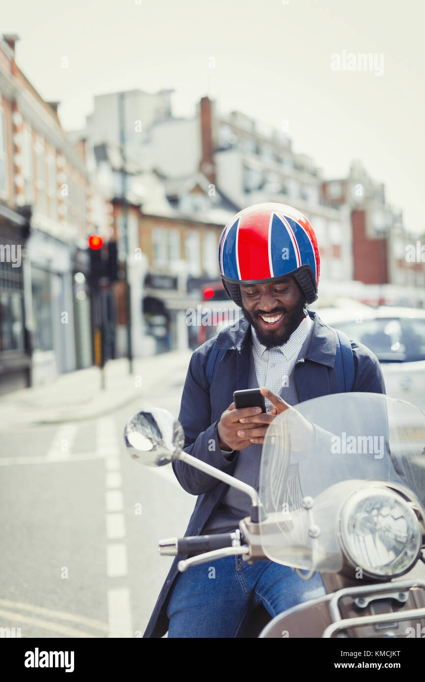 Jeune homme d'affaires souriant dans un casque sur scooter de moteur, texto avec téléphone cellulaire dans la rue urbaine Banque D'Images