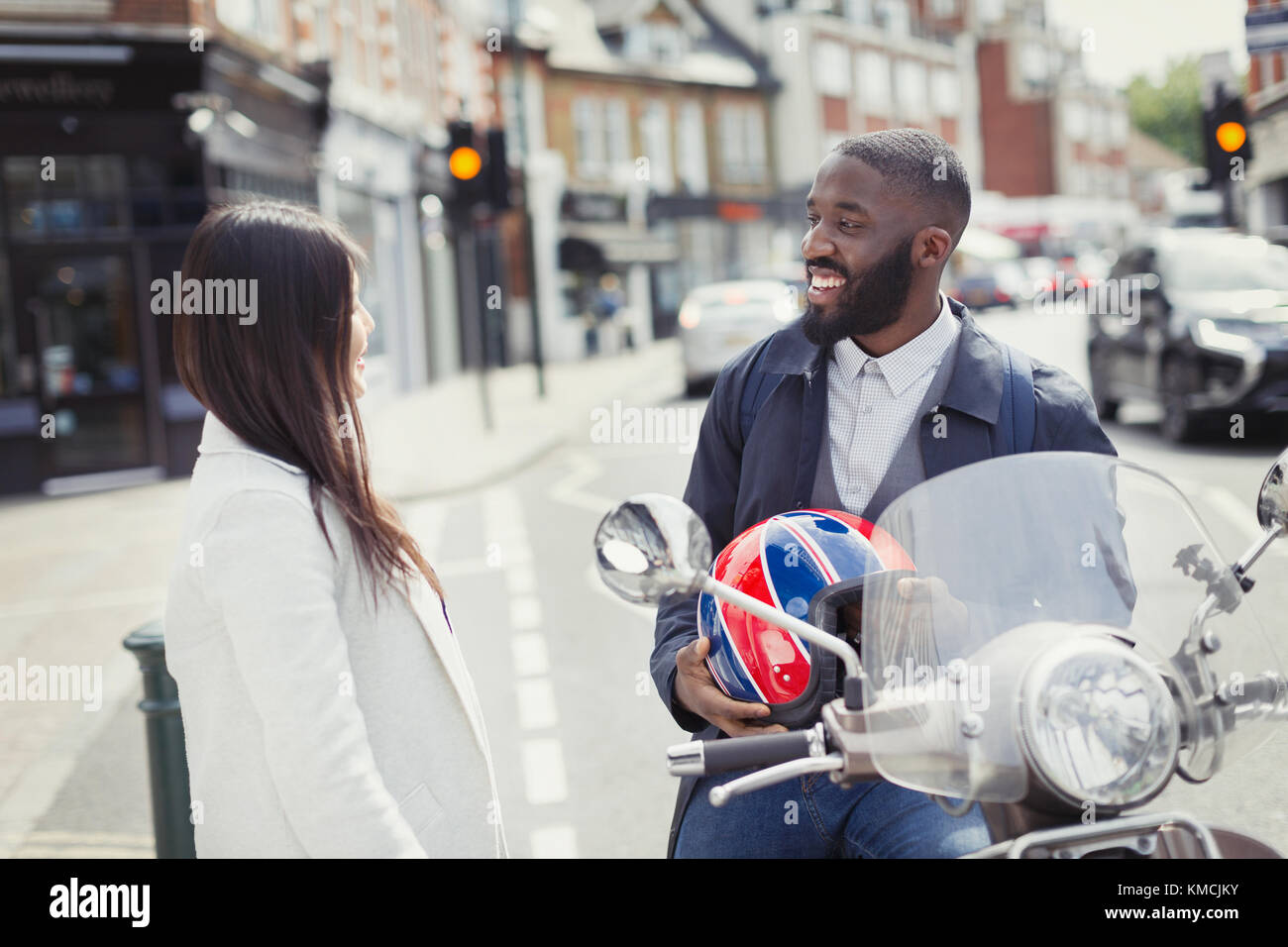 Un jeune homme d'affaires souriant sur un scooter à moteur parle à un ami sur rue urbaine Banque D'Images