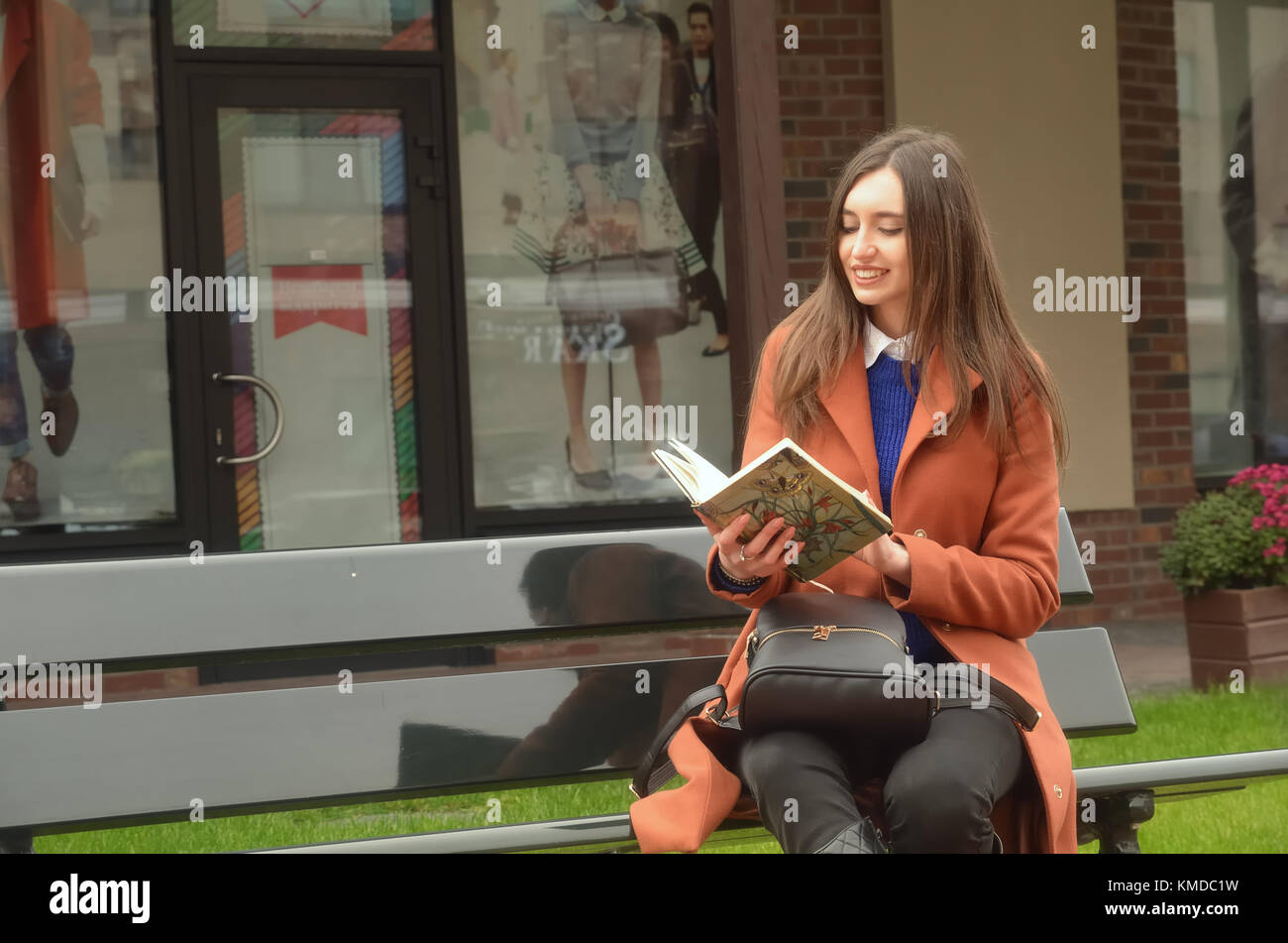 Une belle jeune fille aux cheveux longs assis sur le banc et se balancer un ordinateur portable, en vue de face, derrière les boutiques et d'une pelouse Banque D'Images