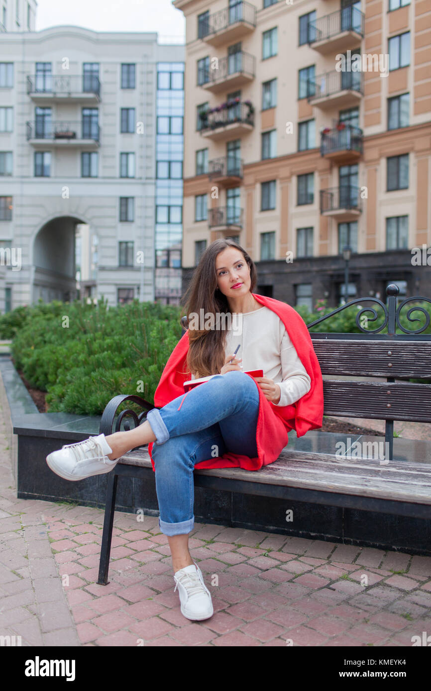 Une fille attrayante avec de longs cheveux bruns et un sourire blanc de dent s'assoit sur un banc et écrit ses pensées sur le fond urbain dans un carnet rouge.Elle Banque D'Images