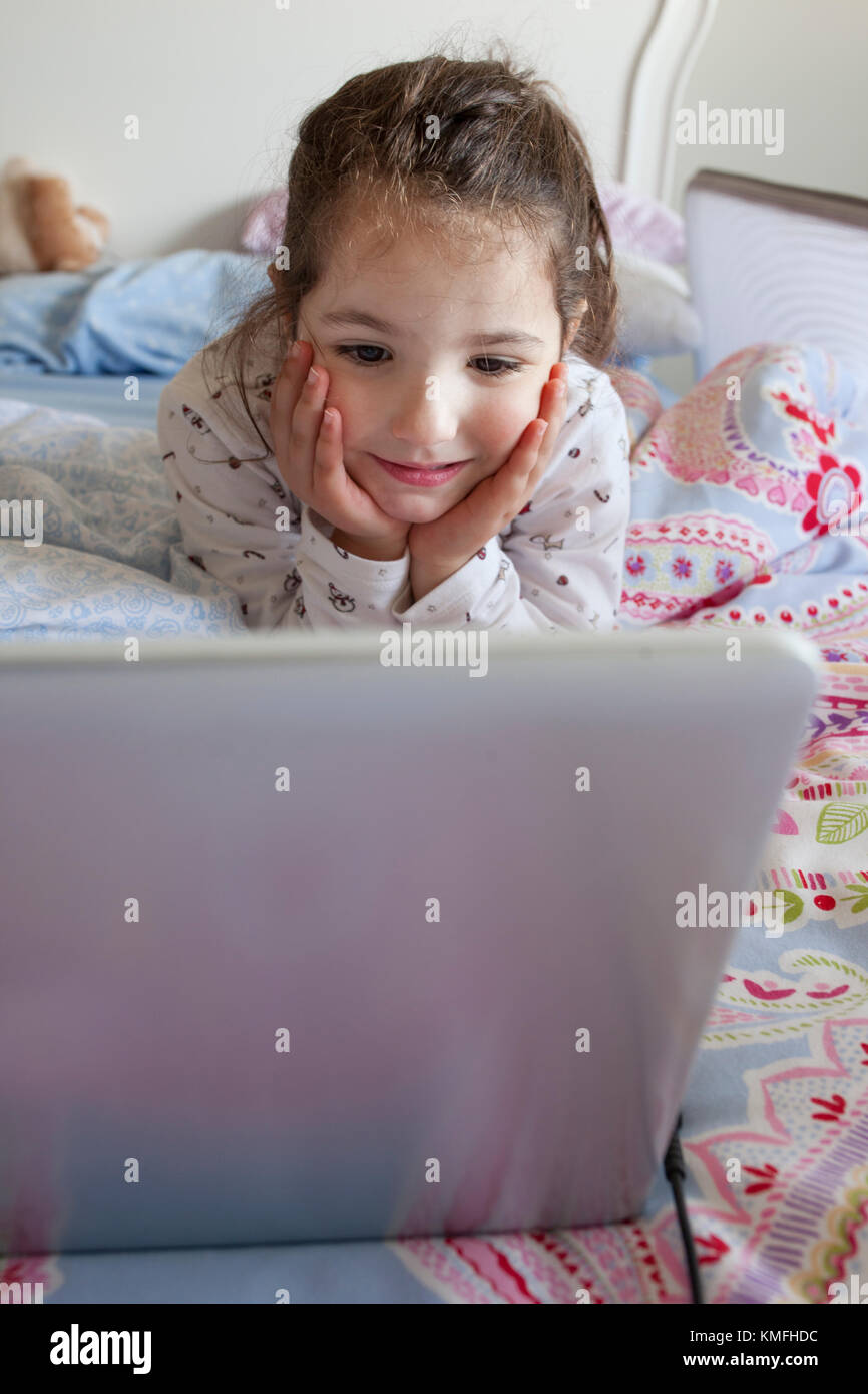 Petite fille au lit et jouer avec un ordinateur portable dans sa chambre. Elle a l'air de s'amuser Banque D'Images