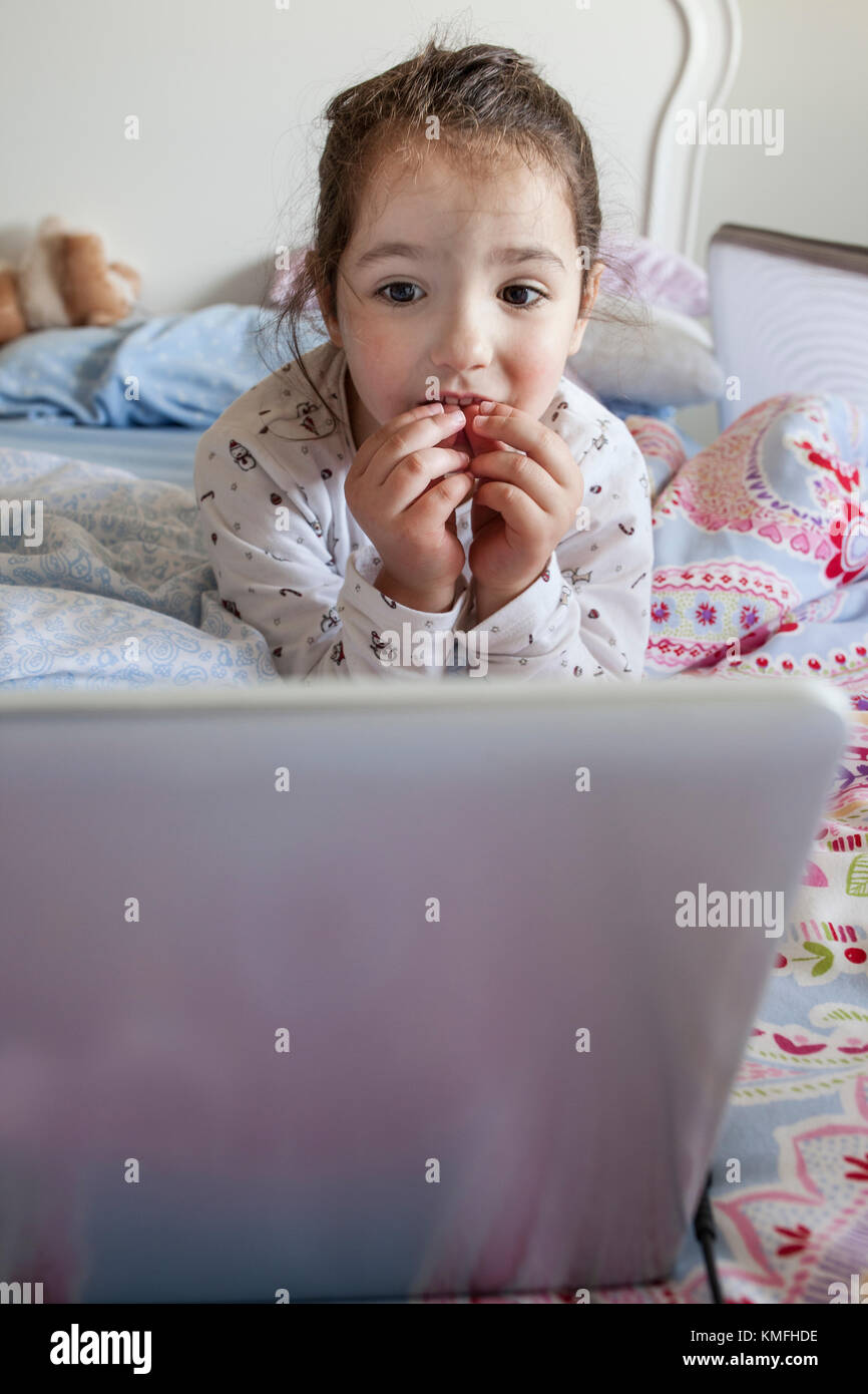 Petite fille au lit et jouer avec un ordinateur portable dans sa chambre. Elle a l'air intrigué Banque D'Images