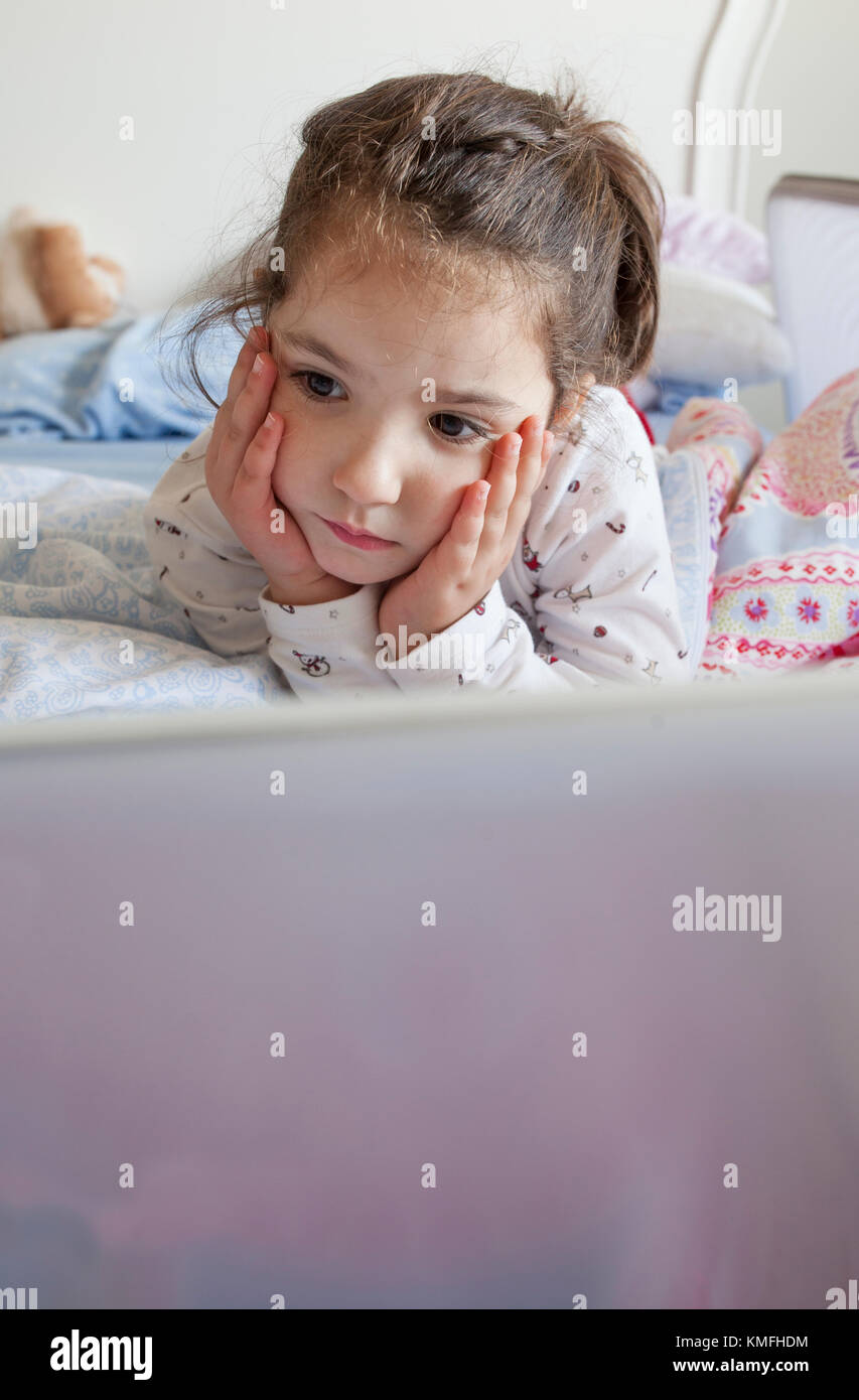 Petite fille couchée au lit et jouant avec un ordinateur portable dans sa chambre.Elle semble inattentive Banque D'Images