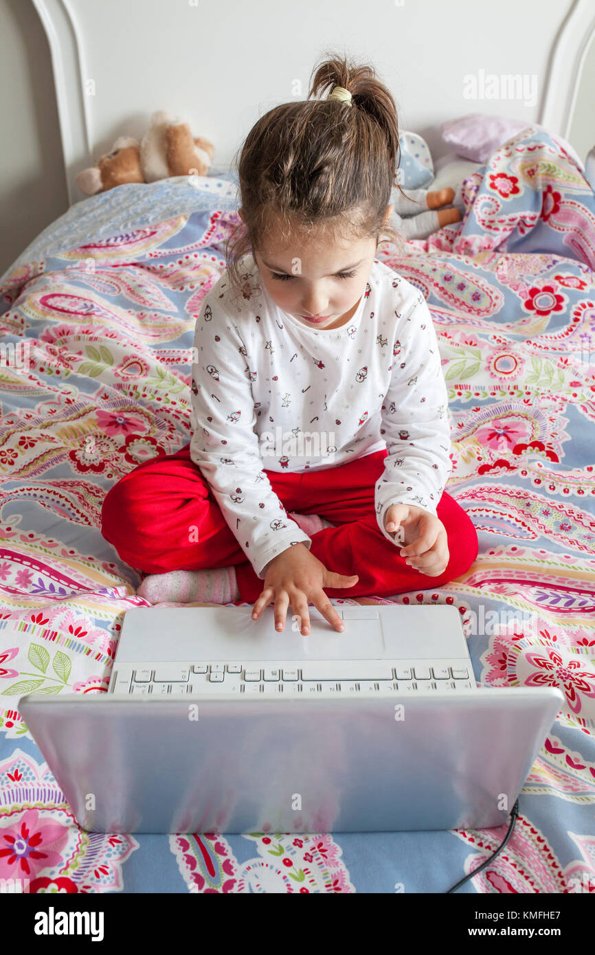 Petite fille assise dans le lit et jouer à des jeux en ligne avec un ordinateur portable Banque D'Images