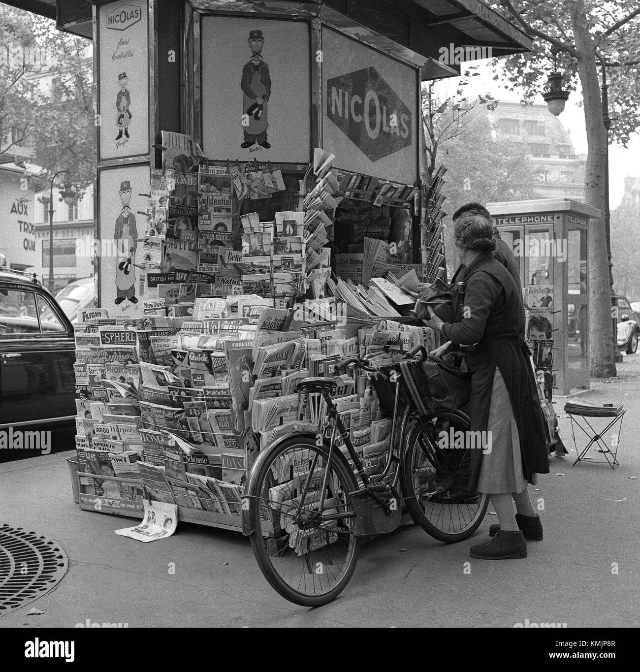 Années 1950, tableau historique des clients à un kiosque à journaux dans le centre de Paris, France avec advertsing pour Nicolas, le parisien établi depuis longtemps wine company. Banque D'Images
