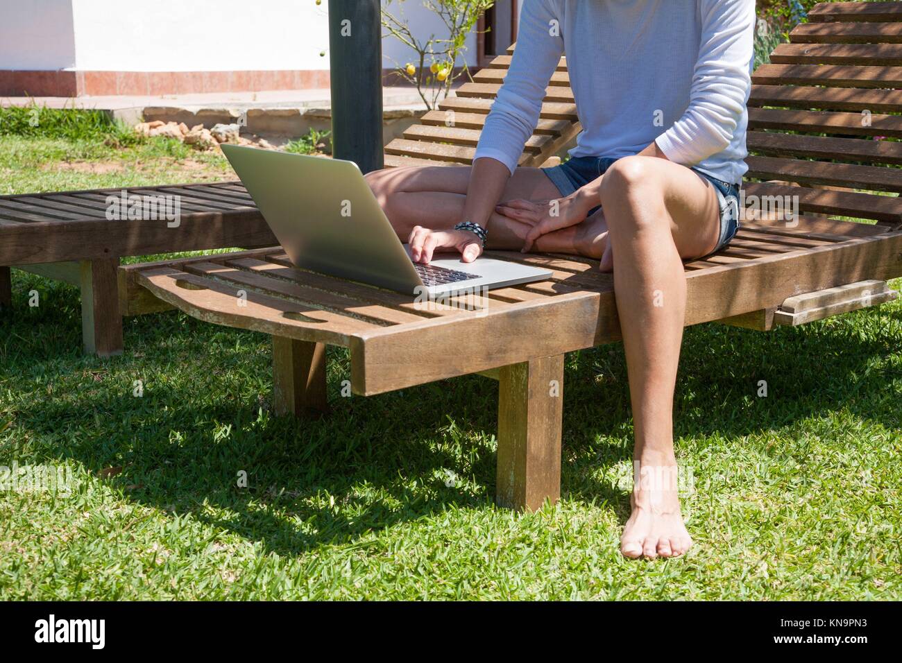 Brunette woman avec chemise blanche et jeans shorts barefoot using laptop  sitting on salon en bois chaise longue à jardin Photo Stock - Alamy