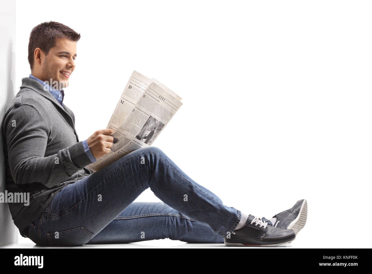 Jeune homme assis sur le plancher de lire le journal et appuyé contre un mur isolé sur fond blanc Banque D'Images