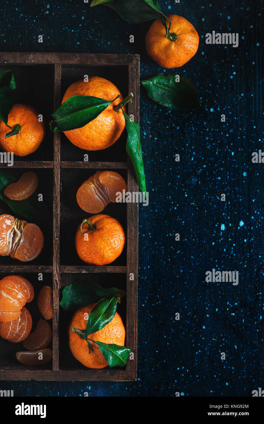 Mandarines dans une caisse de bois sur un fond sombre. Un assortiment de clémentines avec des feuilles vertes. La photographie alimentaire sombre avec des fruits orange vif et Banque D'Images