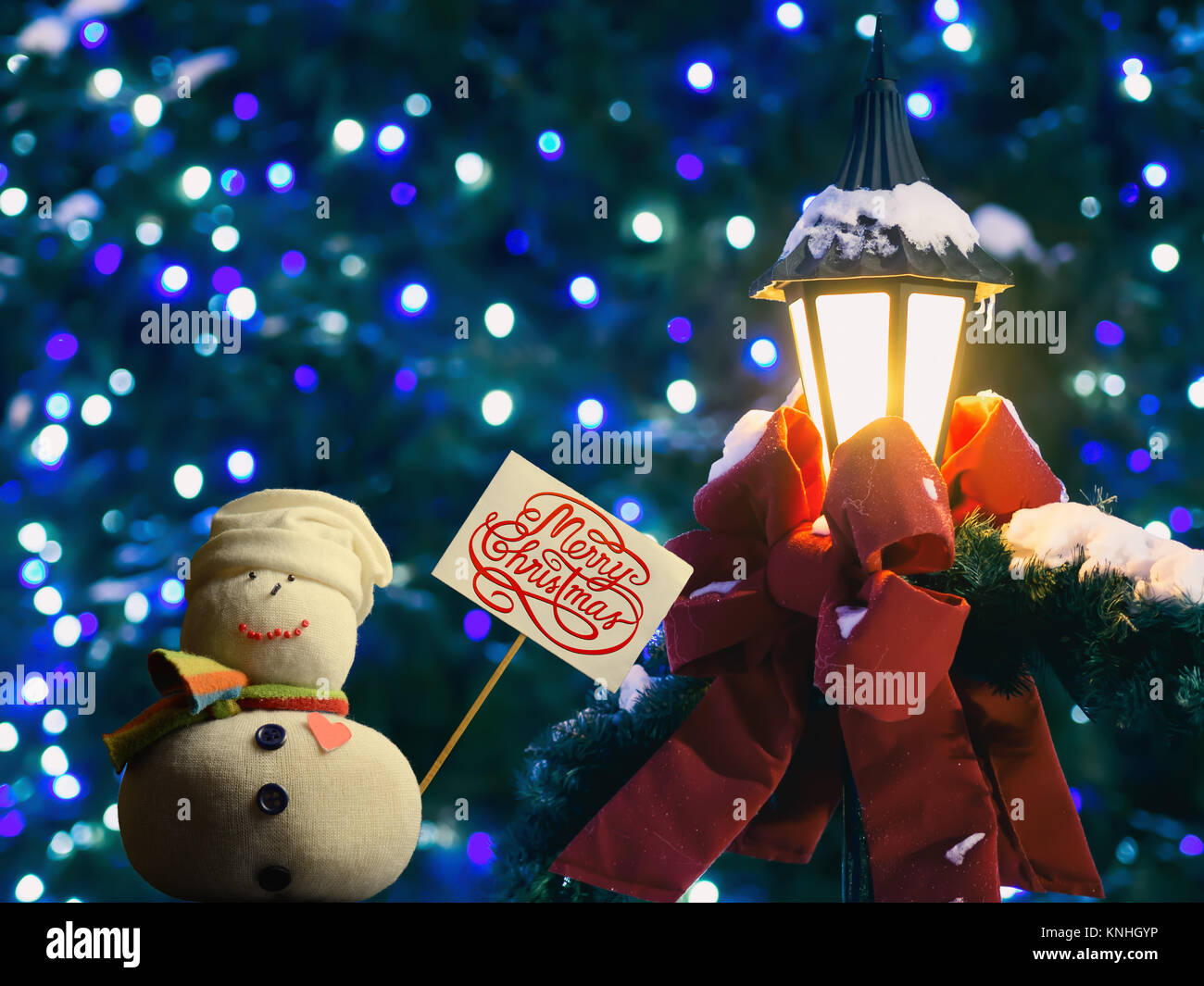 La figure bonhomme holding a placard. Texte "Joyeux Noël !". Lanterne rue enveloppée d'un ruban rouge et des branches de sapin. Beau bokeh. Banque D'Images
