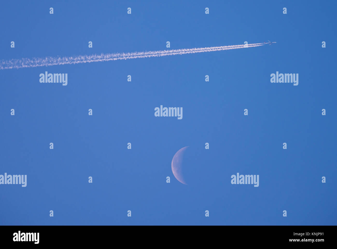 Contrails sur la lune. Avion de ligne à réaction en hauteur créant des traînées de vapeur devant un croissant de lune déclinant dans un ciel bleu jour. Voler haut Banque D'Images