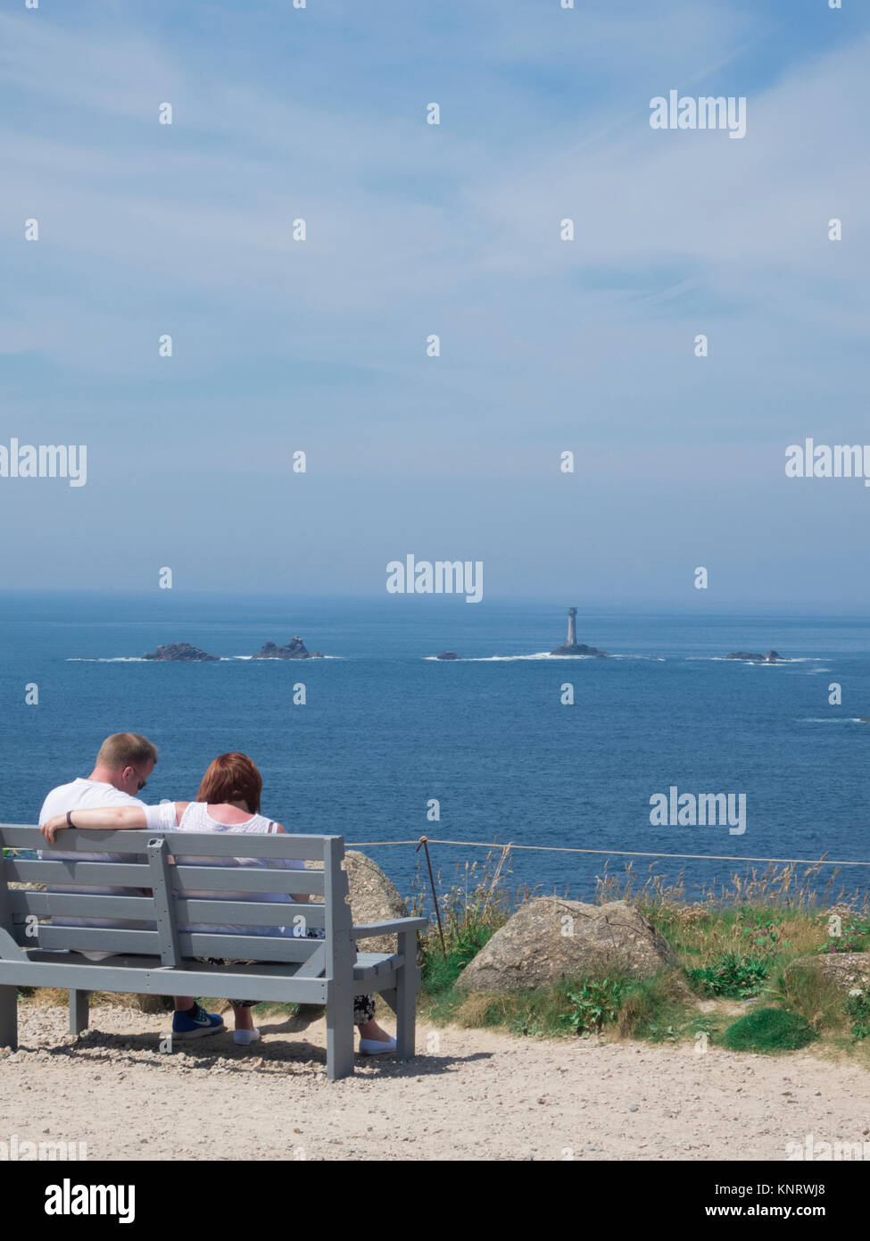 Young Caucasian couple assis sur un banc à Land's End en regardant le phare de Drakkars, Péninsule de Penwith, Cornwall, England, UK en été Banque D'Images