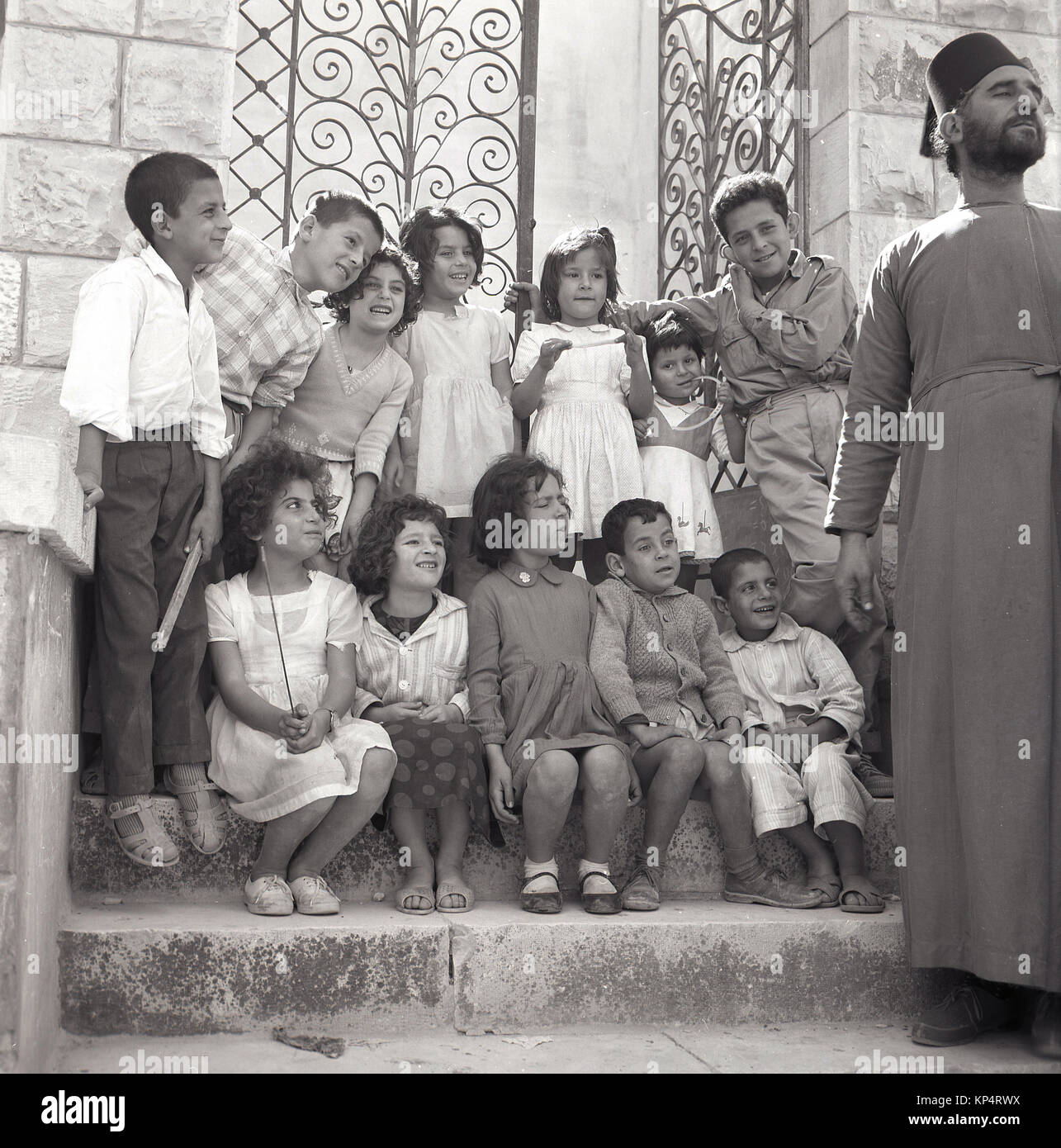 Années 1950, tableau historique, un homme barbu prêtre avec chapeau et la robe a un peu de plaisir avec un groupe d'enfants assis et debout sur les marches à l'extérieur d'une entrée fermée, la Jordanie. Banque D'Images