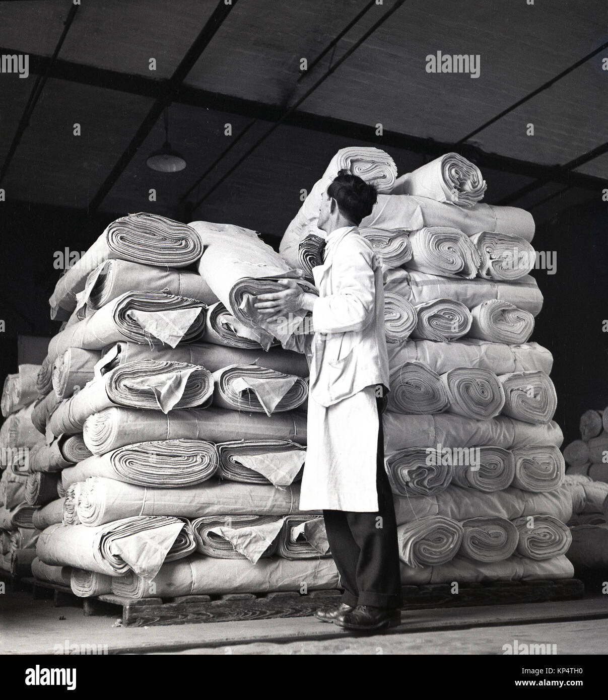 Années 1950, tableau historique, un travailleur en combinaisons blanches contrôler un rouleau de tissu en lin parmi les nombreux qui sont soigneusement empilés sur des palettes, en Irlande du Nord. Banque D'Images