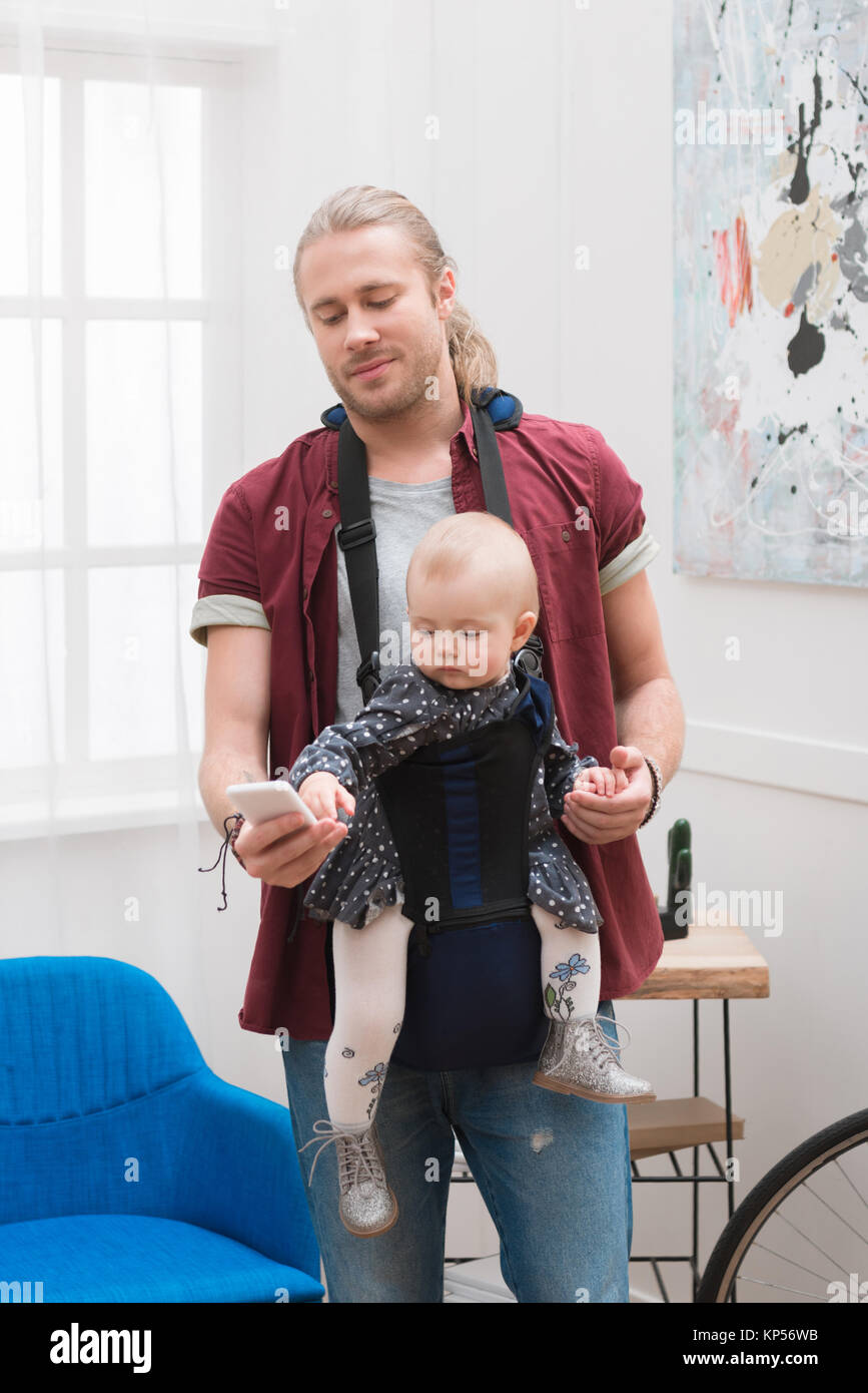 Père debout avec fils en porte-bébé et looking at smartphone Banque D'Images