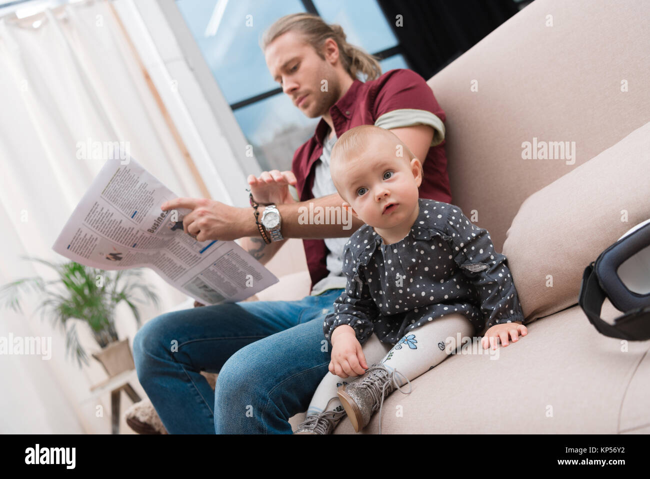 Little baby girl sitting on sofa tout en lisant le journal de son père à la maison Banque D'Images