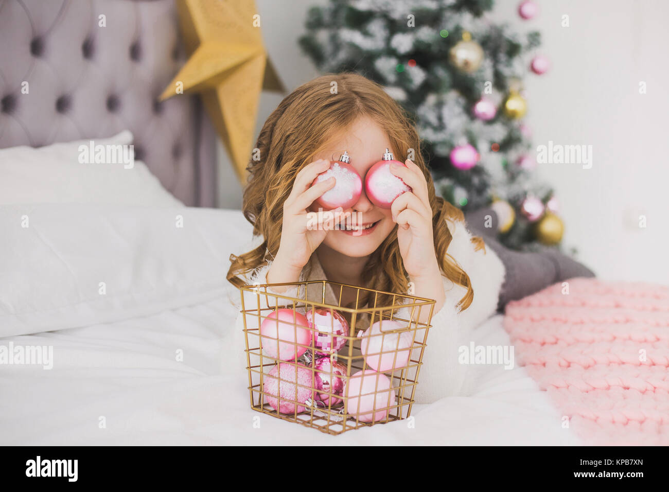 Portrait of cute little girl le matin de Noël dans votre intérieur. Enfant payant avec des décorations de Noël, faisant de l'œil tout en jetant des boules rose Banque D'Images